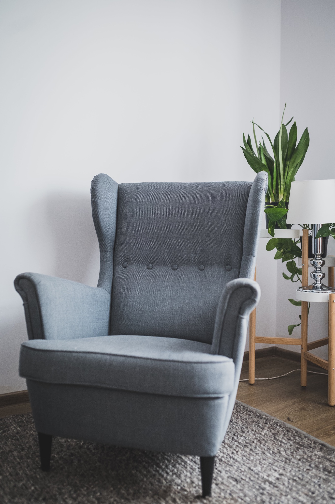 Szary, tapicerowany fotel typu uszak z trzema guzikami na oparciu, stojący na szarym dywanie. Obok stoi stolik z drewnianymi nogami, na którym znajduje się roślina doniczkowa i lampka.