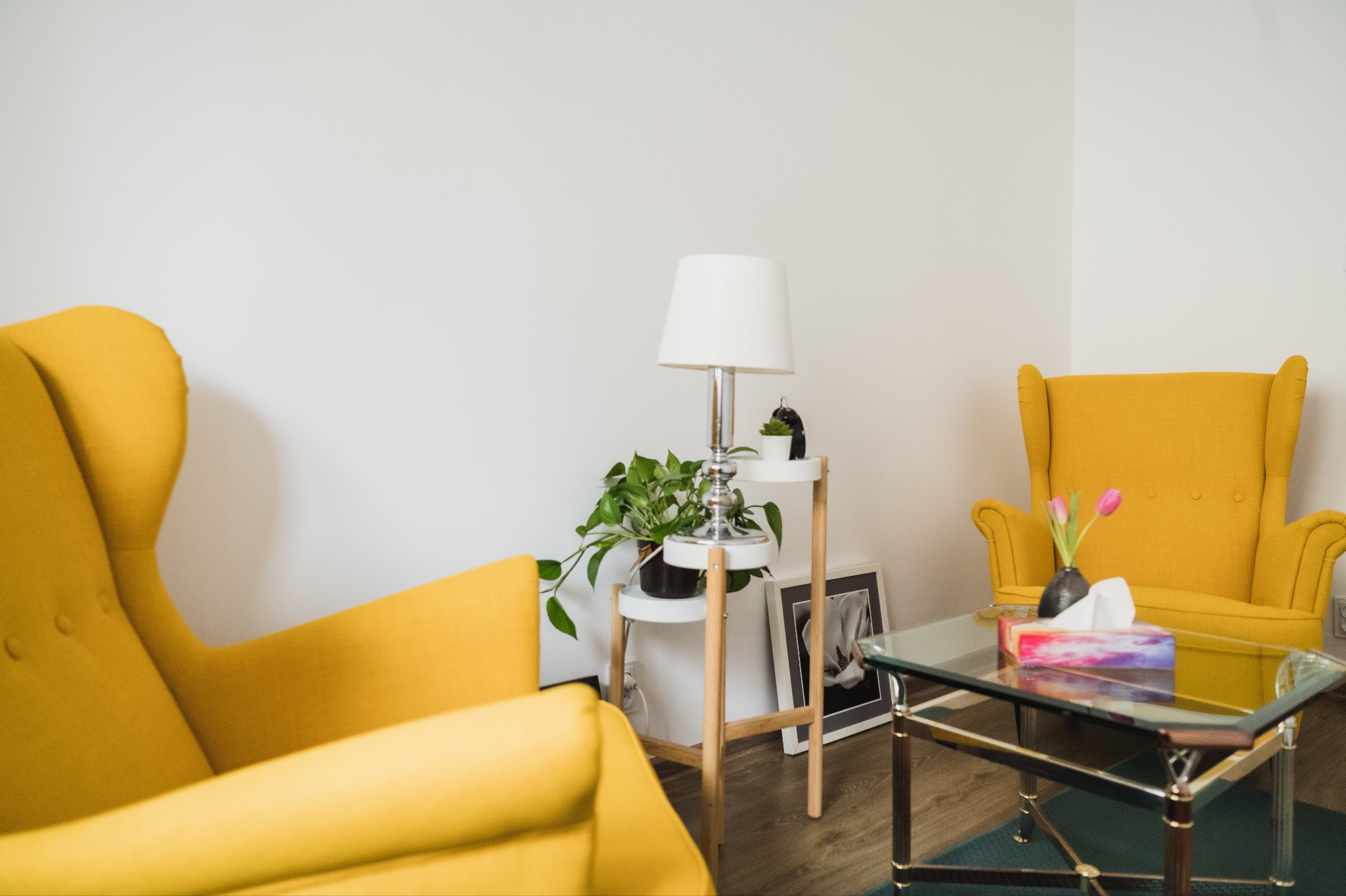 Ustawienie pokoju psychoterapeuty: dwa żółte fotele uszakowe, szklany stolik kawowy z kwiatami i pudełkiem chusteczek, lampa na stoliku pomocniczym z rośliną doniczkową i czarno-białe zdjęcie w ramce.
