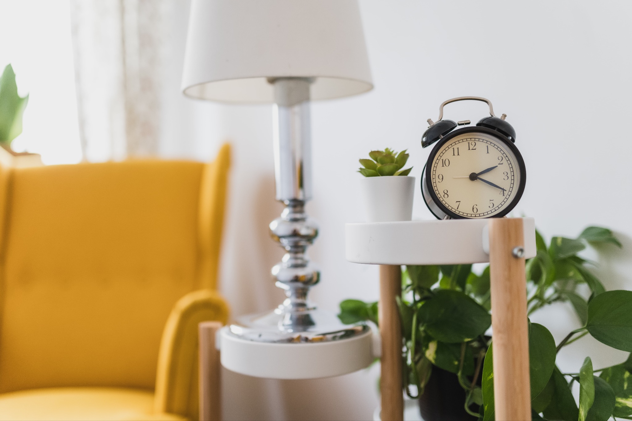 Czarny budzik ustawiony na małym, białym stoliku obok rośliny doniczkowej i lampy w pokoju w Krakowie, z fragmentem żółtego fotela w tle.
