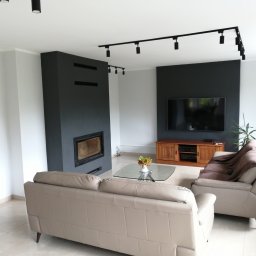 AgMil usługi remontowo-budowlane - Jasny salon z beżową sofą, szarą ścianą z kominkiem, drewnianą szafką RTV i czarnym oświetleniem punktowym na szynach.