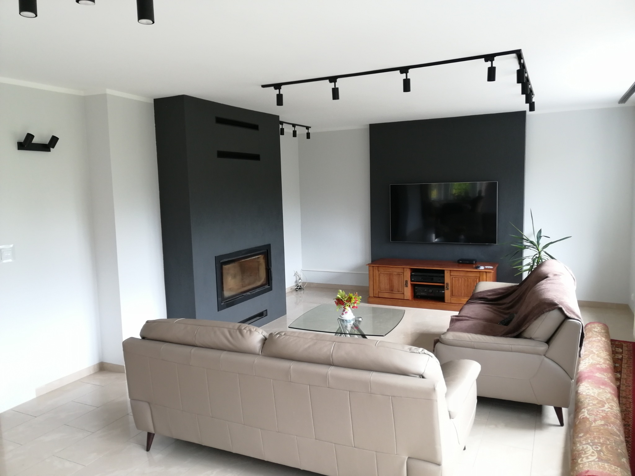 Jasny salon z beżową sofą, szarą ścianą z kominkiem, drewnianą szafką RTV i czarnym oświetleniem punktowym na szynach.