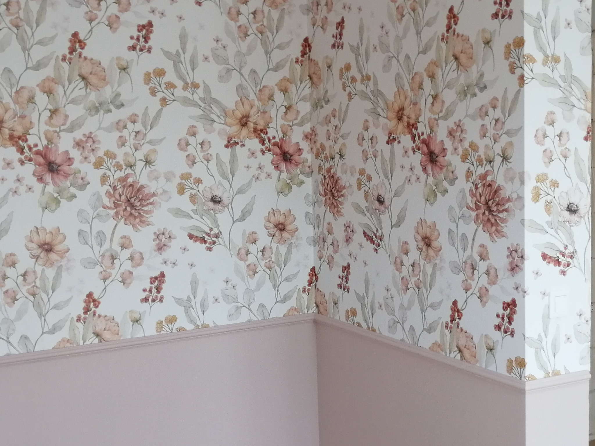 Ściana z delikatnym, kwiatowym wzorem tapety w pastelowych odcieniach różu i beżu, połączona z dolną częścią pomalowaną na jednolity, pudrowy róż, tworząc efekt dekoracyjny.