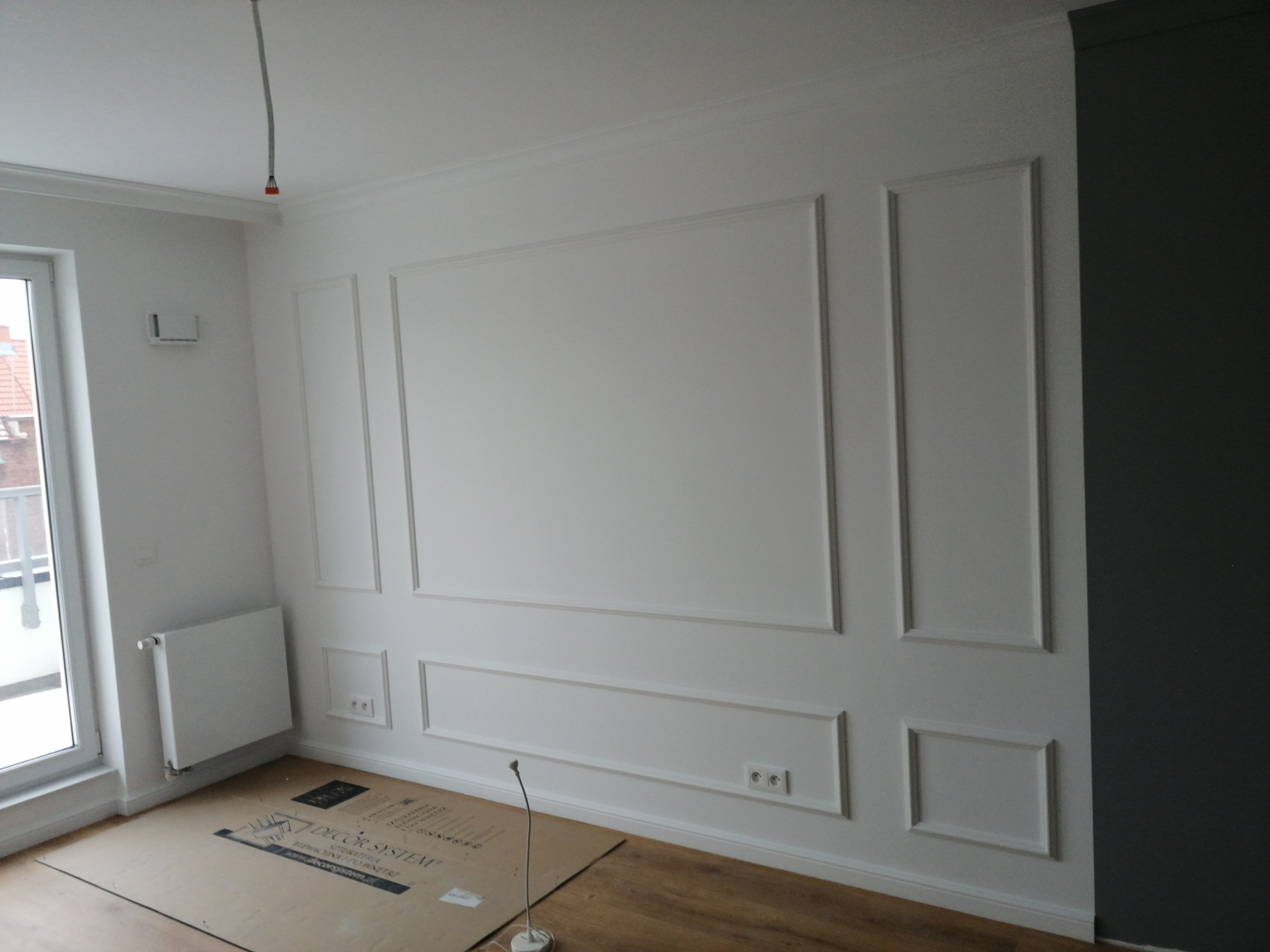 Ściana z dekoracyjnymi listwami w trakcie prac, zabezpieczona kartonem na podłodze, widoczne gniazdka elektryczne i grzejnik.