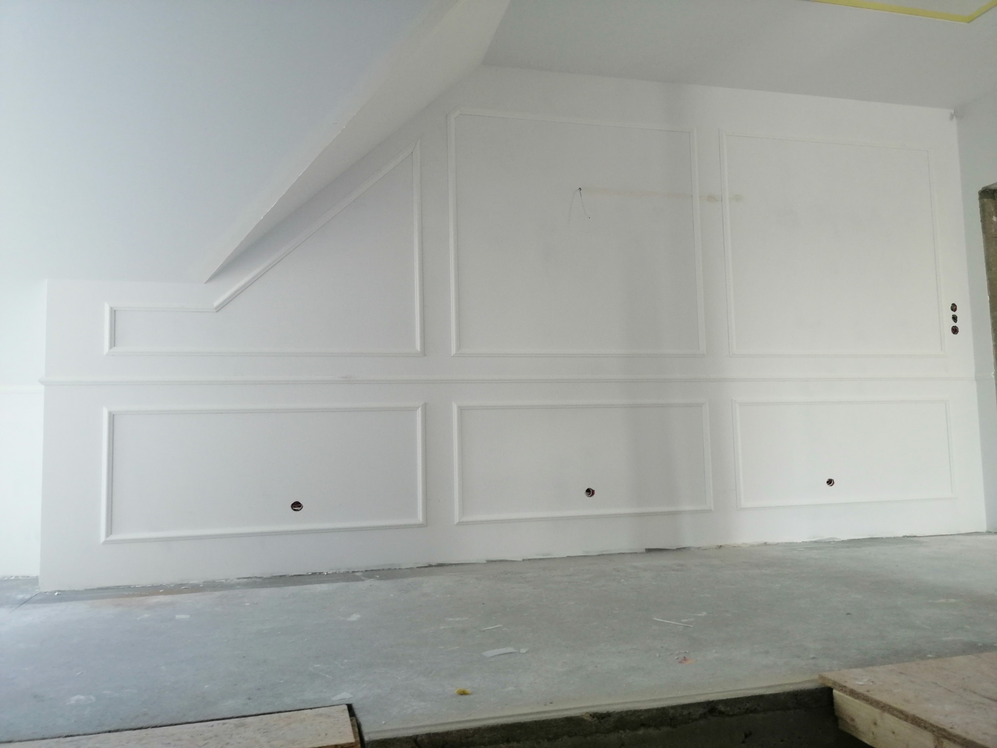 Ściana z białymi panelami dekoracyjnymi w kształcie prostokątów i kwadratów, widoczne otwory na gniazdka elektryczne, surowa podłoga w szarym kolorze, fragment skośnego sufitu.