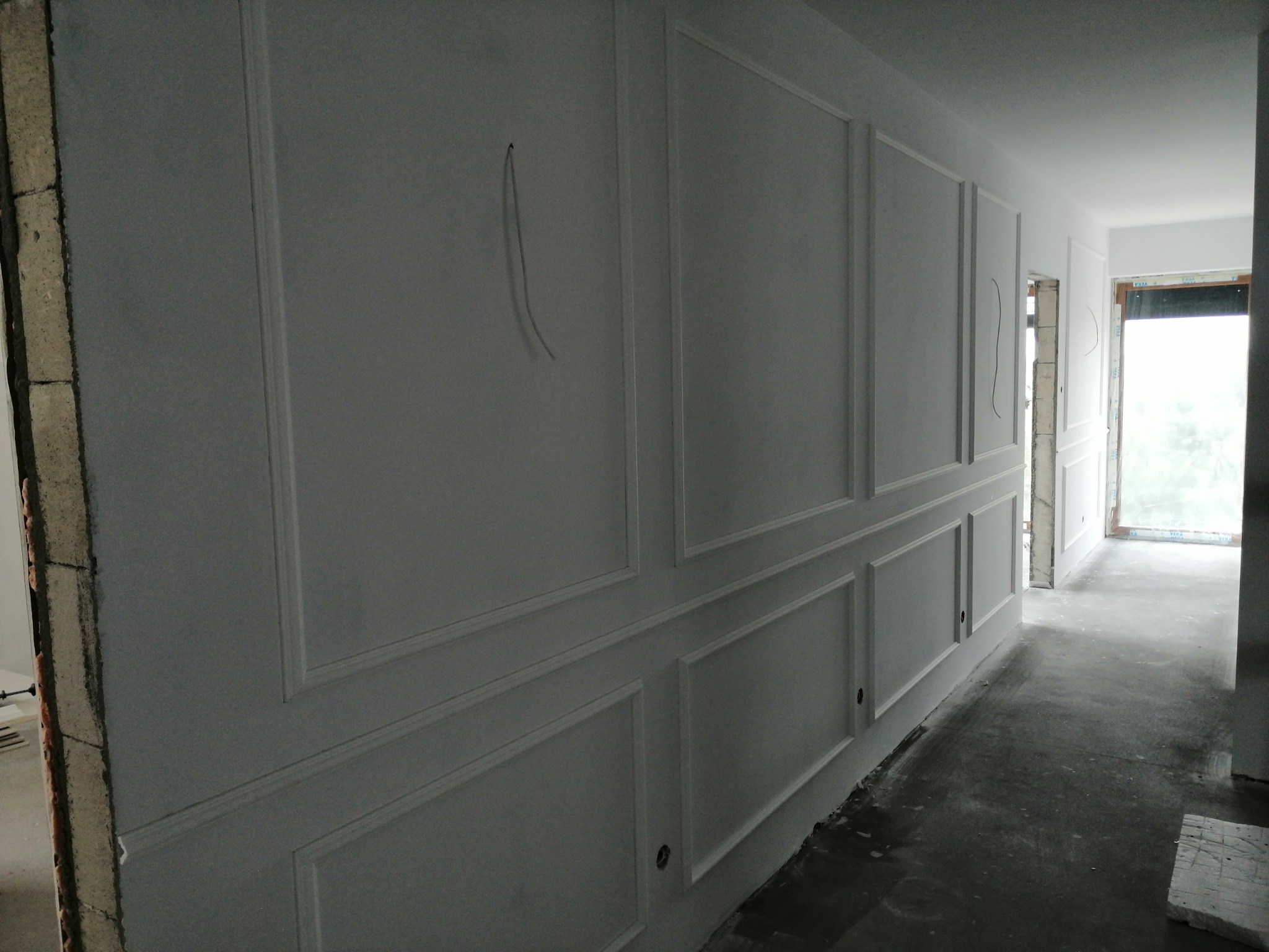 Ściana z białymi panelami dekoracyjnymi w trakcie montażu, widoczne kable elektryczne wystające ze ściany oraz surowy beton przy otworze drzwiowym.