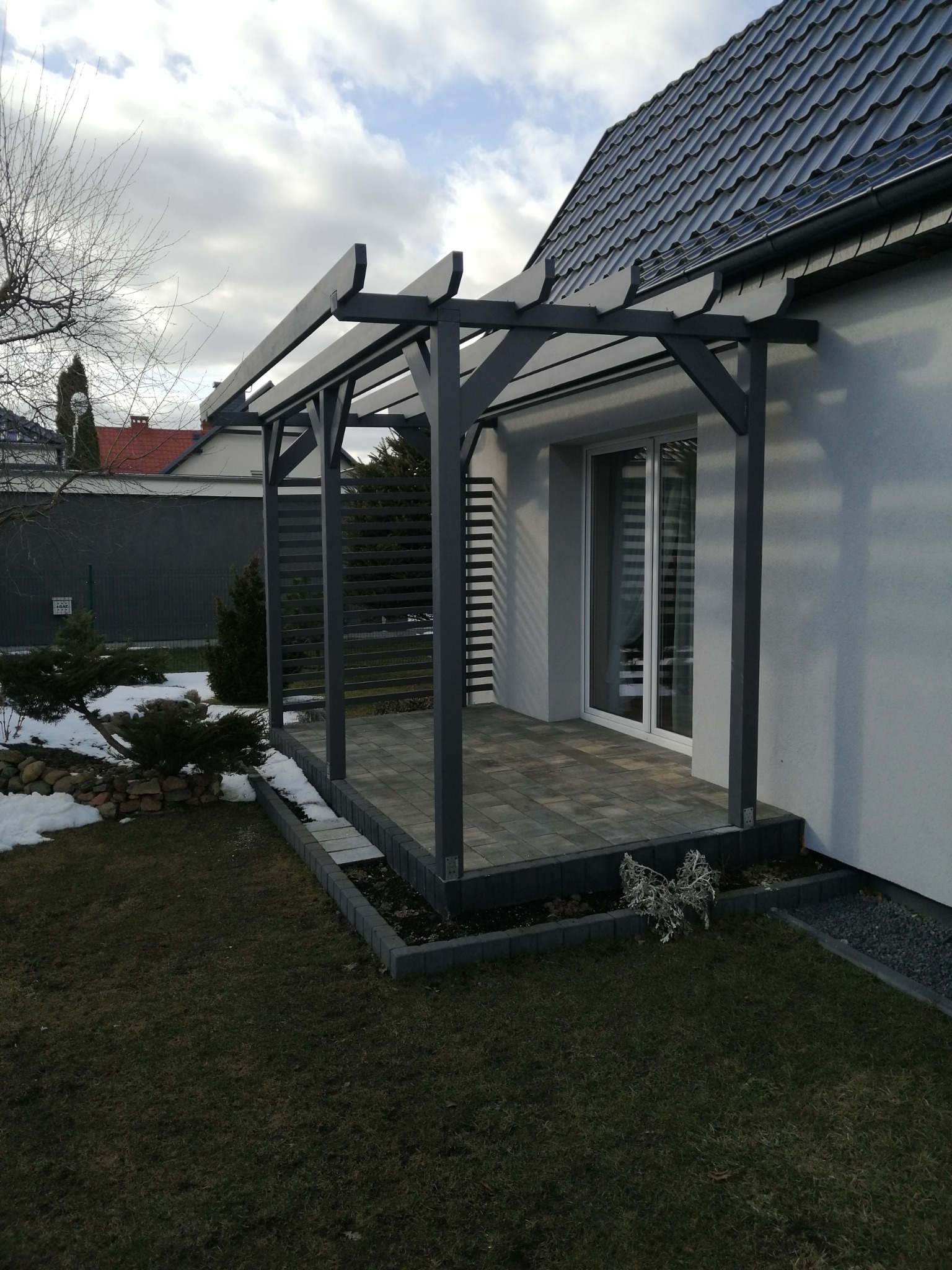 Nowoczesna, szara pergola przylegająca do białego domu z szarym dachem, wyłożona kostką brukową, widoczny fragment ogrodu z resztkami śniegu.