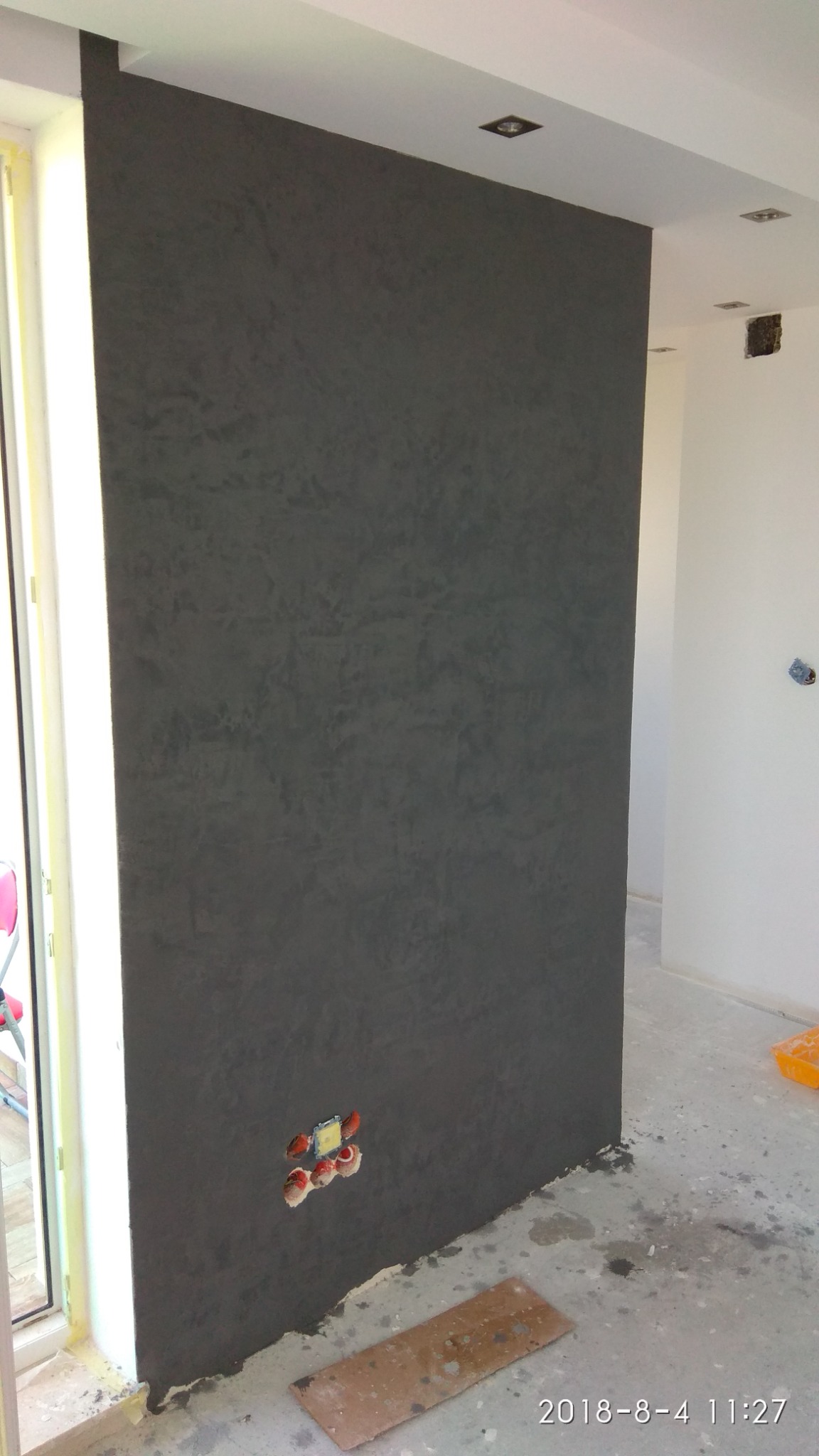 Ściana z dekoracyjną, ciemnoszarą strukturą tynku w trakcie prac wykończeniowych, widoczne gniazdka elektryczne bez osprzętu, fragment podłogi z resztkami zaprawy i płyta.