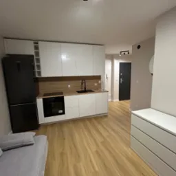 Nowoczesna kuchnia z białymi szafkami, drewnianym blatem i czarną lodówką Amica. Jasne panele podłogowe i szara sofa w rogu. Minimalistyczny design.