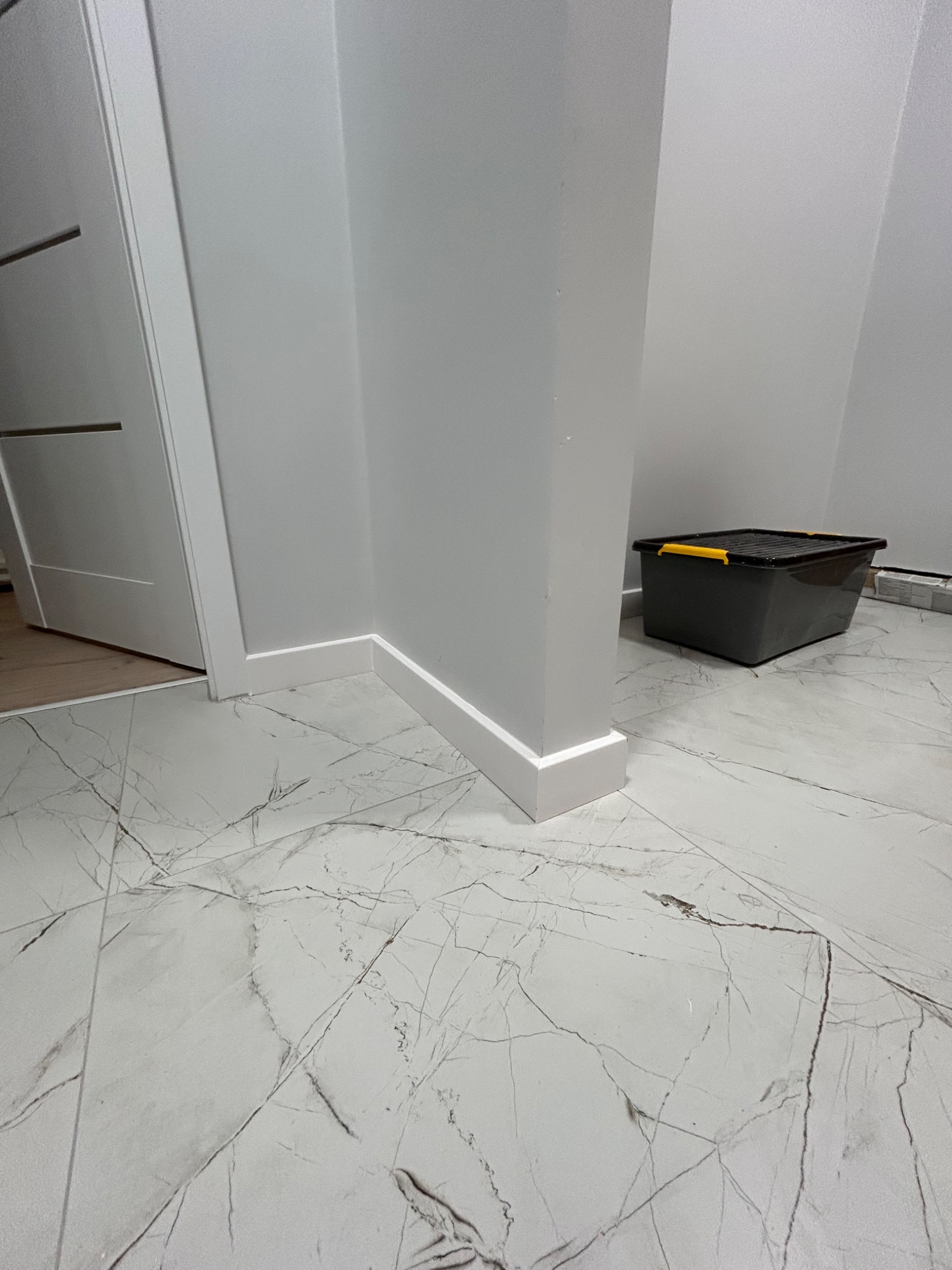 Świeżo pomalowany róg pomieszczenia z białą listwą przypodłogową, widoczny fragment białych drzwi i szare pudełko z żółtymi akcentami na marmurowej podłodze.