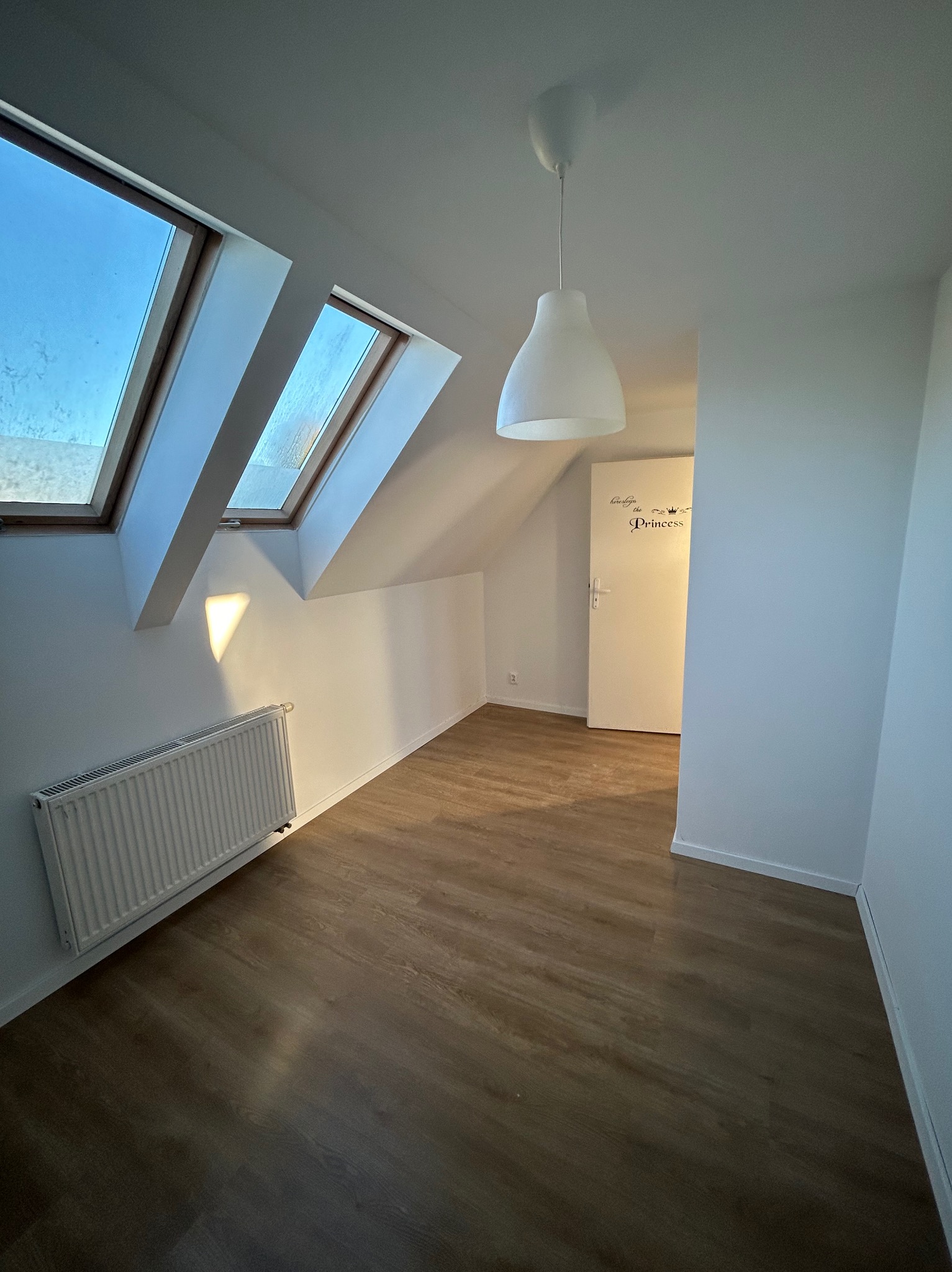 Poddasze po remoncie: drewnopodobne panele, białe ściany z grzejnikiem, okna dachowe i biała lampa wisząca, drzwi z napisem 'Princess'.