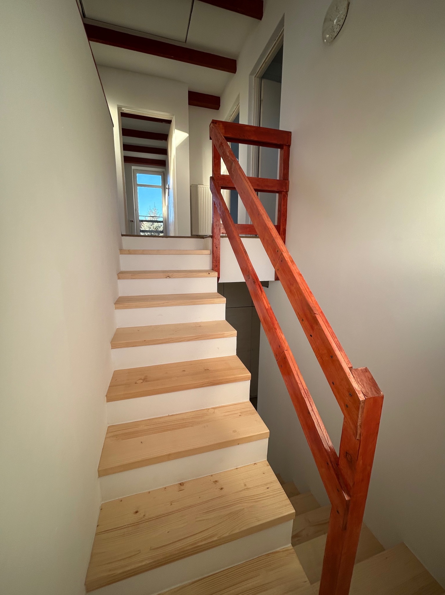 Schody drewniane z pomalowanymi na biało podstopnicami i czerwoną barierką, prowadzące do góry w jasnym wnętrzu.