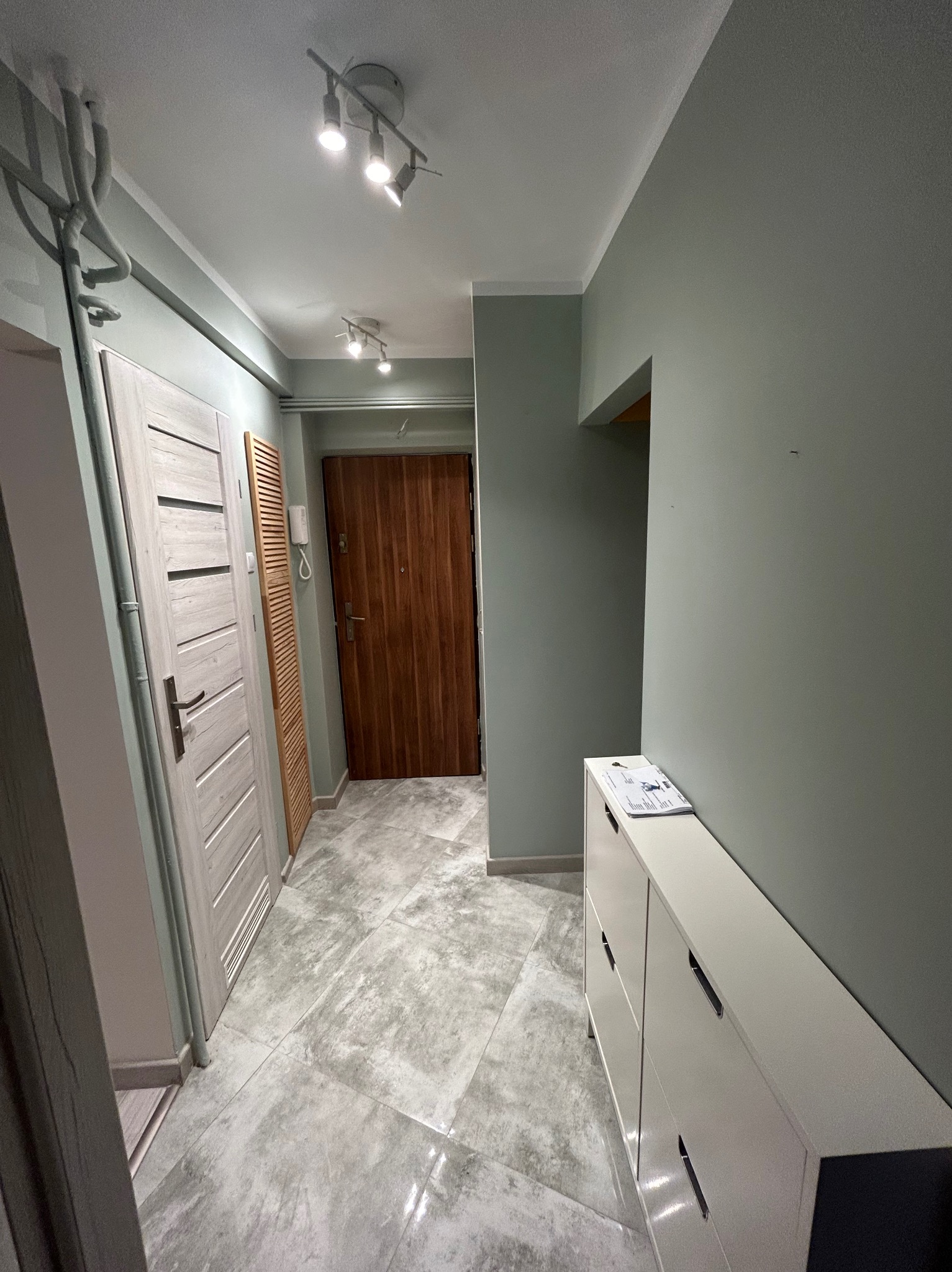 Wnętrze przedpokoju po remoncie: szare płytki podłogowe imitujące marmur, świeżo pomalowane ściany na kolor pistacjowy, biała szafka na buty i drewniane drzwi wejściowe.