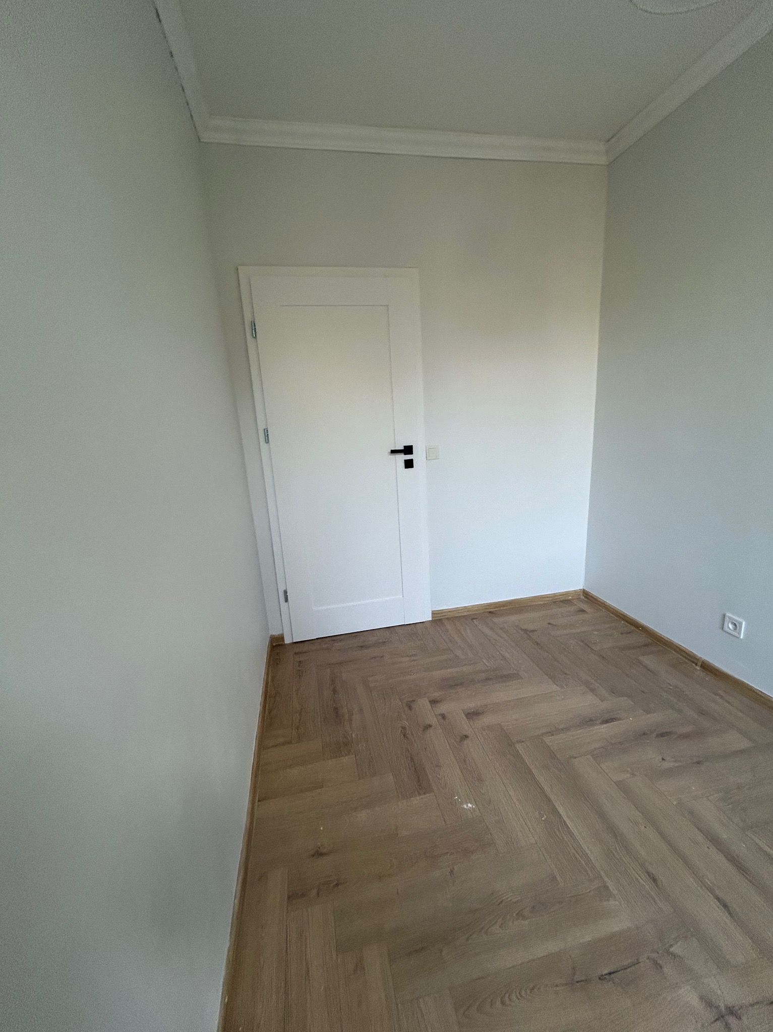Wnętrze pokoju po remoncie: białe ściany, jasna podłoga ułożona w jodełkę, białe drzwi z czarną klamką, listwy przypodłogowe. Widoczne gniazdko elektryczne na ścianie.