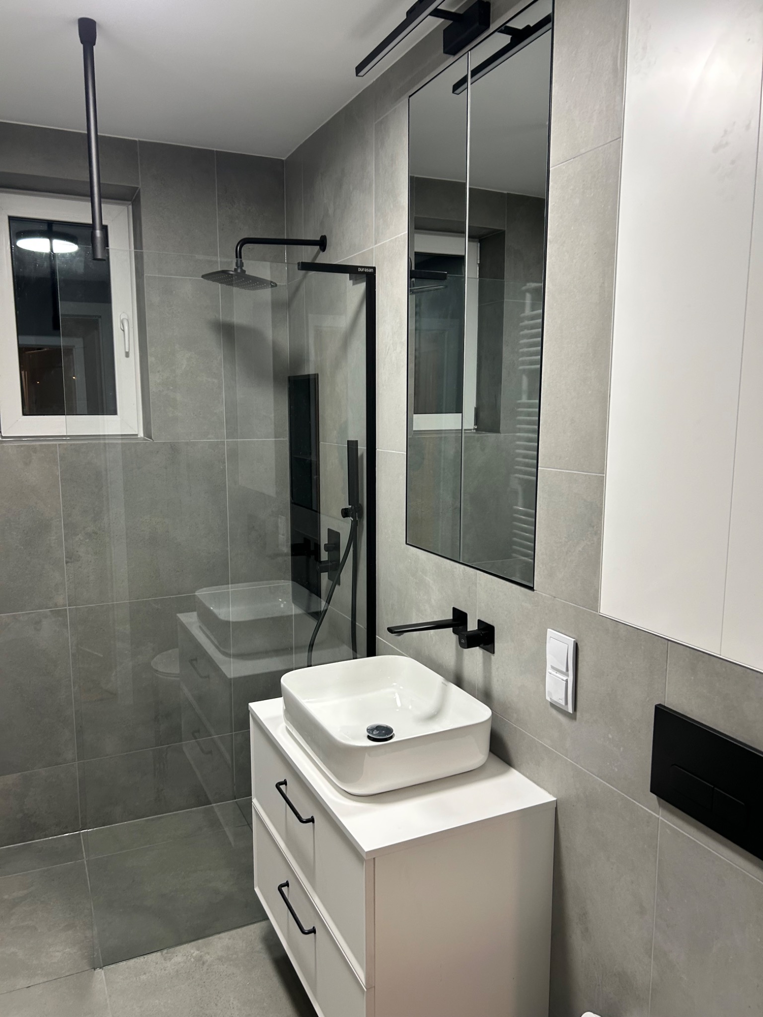 Nowoczesna łazienka z szarymi płytkami, białą szafką pod umywalką i czarną armaturą. Kabina prysznicowa typu walk-in z deszczownicą. Minimalistyczny design.