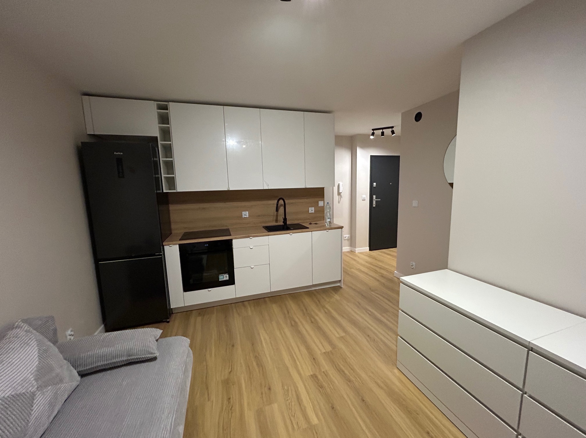 Nowoczesna kuchnia z białymi szafkami, drewnianym blatem i czarną lodówką Amica. Jasne panele podłogowe i szara sofa w rogu. Minimalistyczny design.