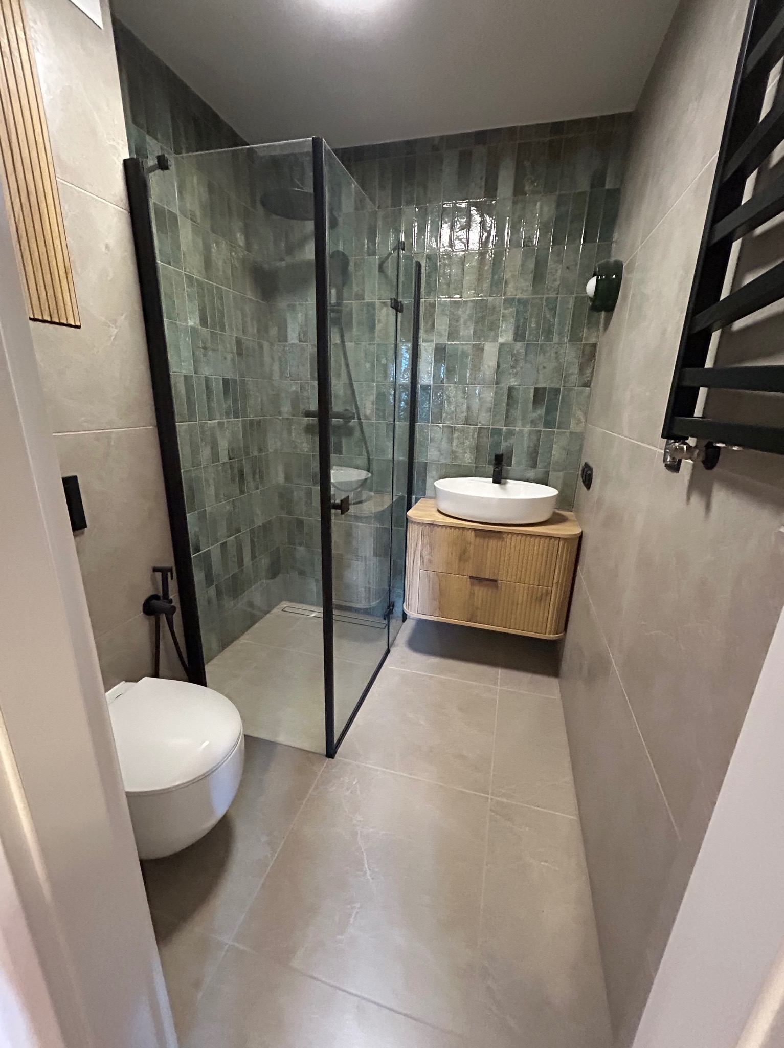 Nowoczesna łazienka z kabiną prysznicową typu walk-in, umywalką nablatową na drewnianej szafce i grzejnikiem drabinkowym. Ściany wyłożone płytkami w odcieniach szarości i zieleni.