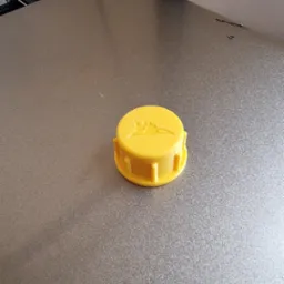 Żółta, okrągła nakładka z wytłoczonym logo na szarej powierzchni drukarki 3D.