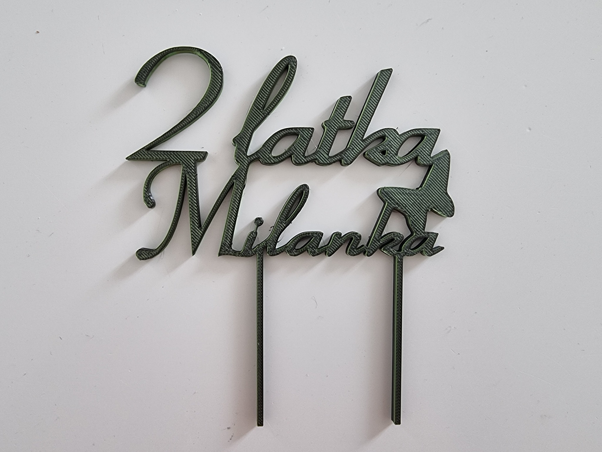 Dekoracja tortu z napisem '2 latka Milanka' wykonana z zielonego tworzywa sztucznego, leżąca na białym tle.