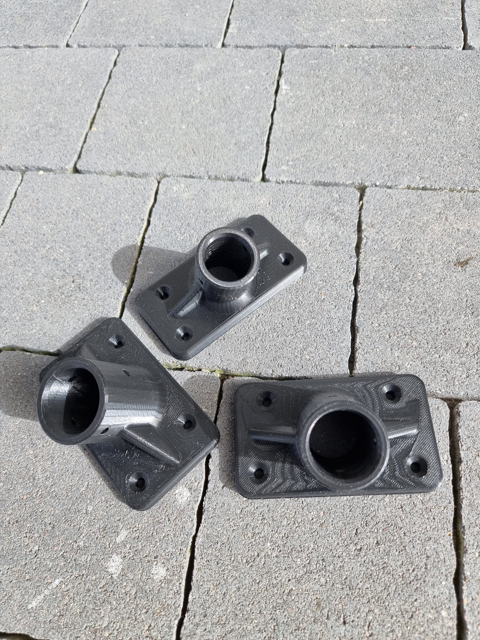 Trzy czarne, plastikowe uchwyty rurowe z otworami na śruby, wydrukowane w technologii 3D, leżące na szarej kostce brukowej w słoneczny dzień.