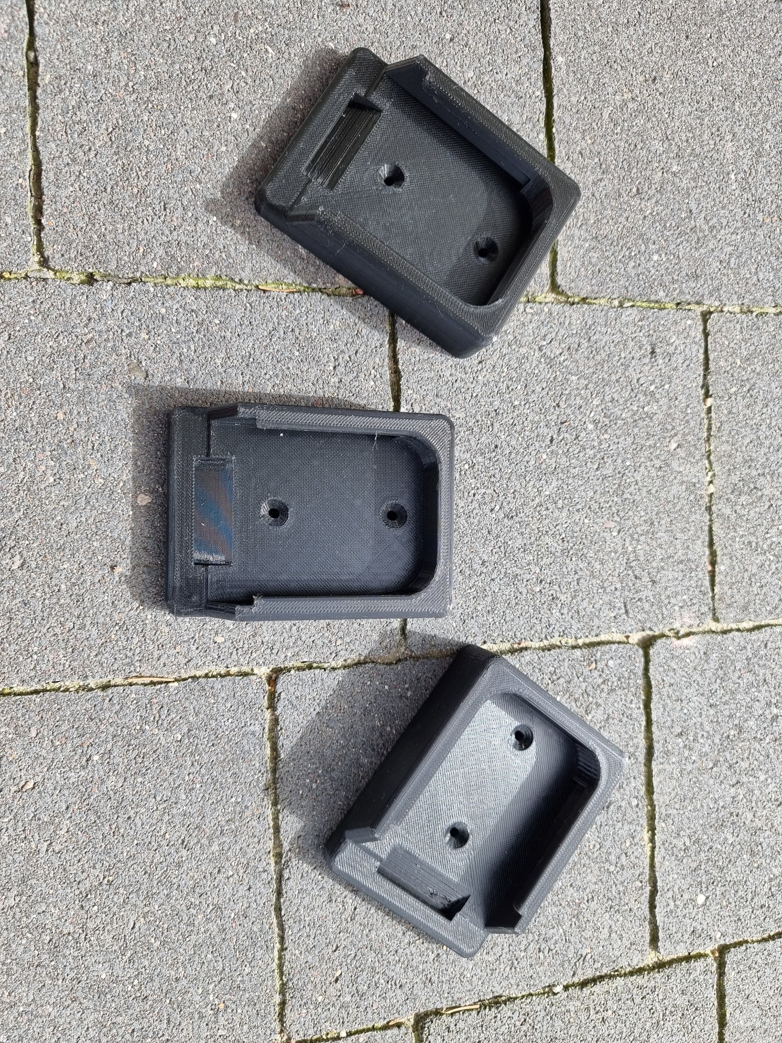 Trzy czarne, plastikowe elementy montażowe z widoczną strukturą druku 3D, leżące na szarej kostce brukowej w słoneczny dzień.