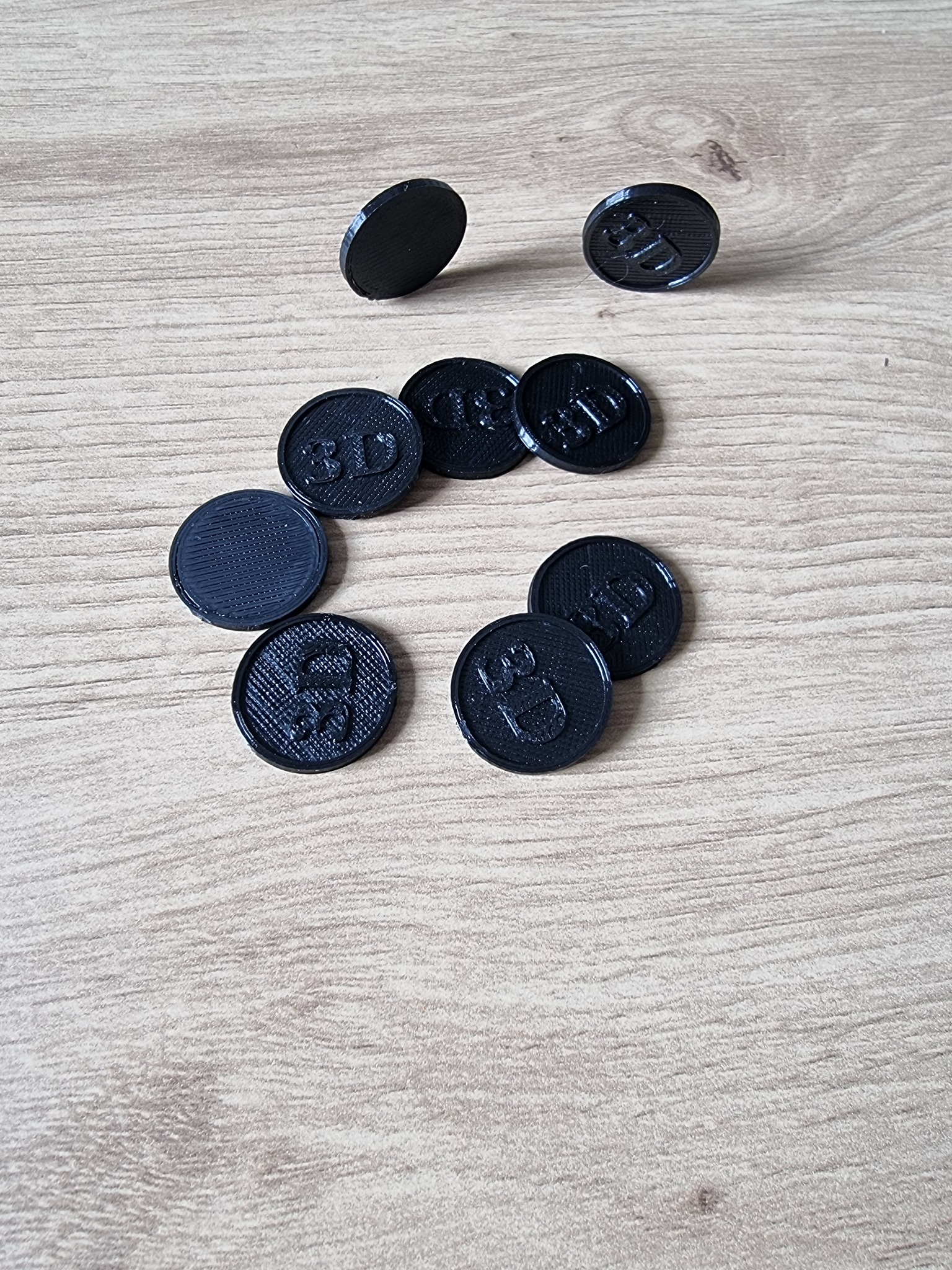 Czarne, okrągłe przedmioty z nadrukiem 3D na drewnianym tle, jeden przedmiot stoi pionowo.