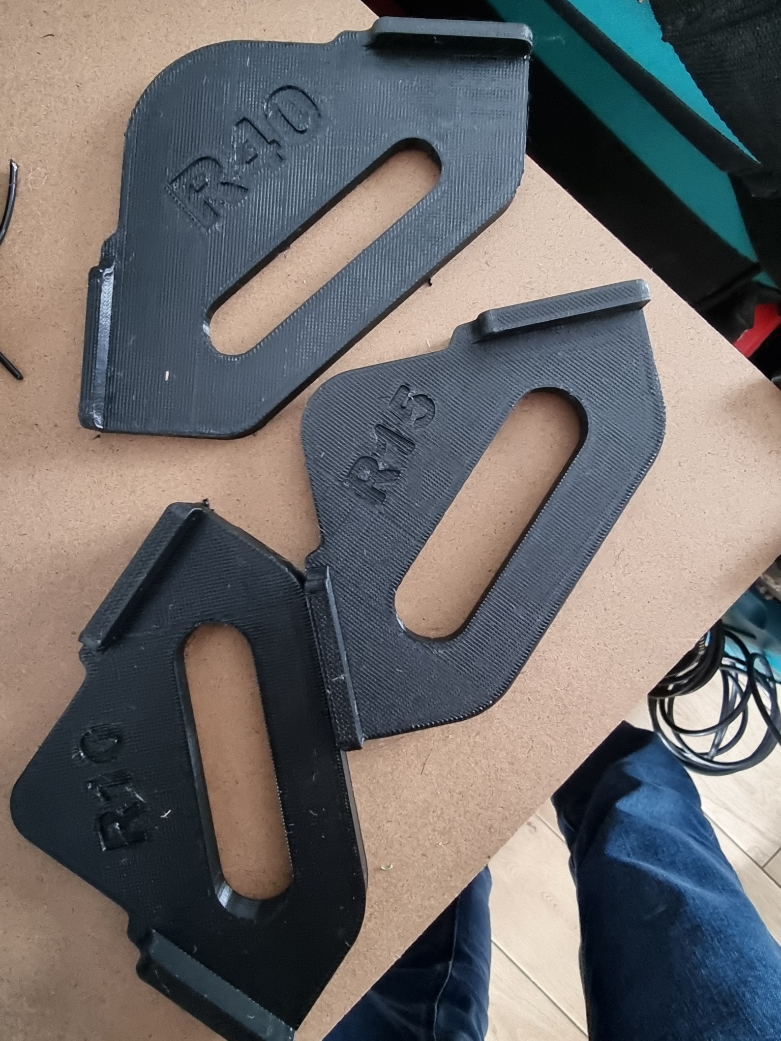 Trzy czarne, plastikowe elementy z nadrukowanymi oznaczeniami R40 i R15, wykonane techniką druku 3D, leżące na brązowej płycie w warsztacie.