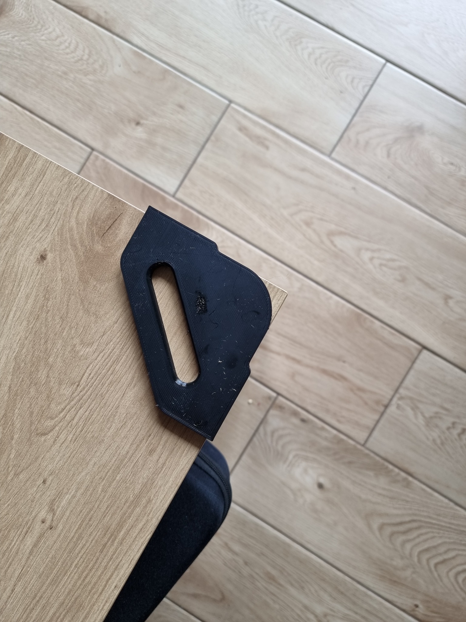 Przycięty róg płyty drewnopodobnej za pomocą czarnego, plastikowego narzędzia z podłużnym otworem, na tle podłogi z paneli.