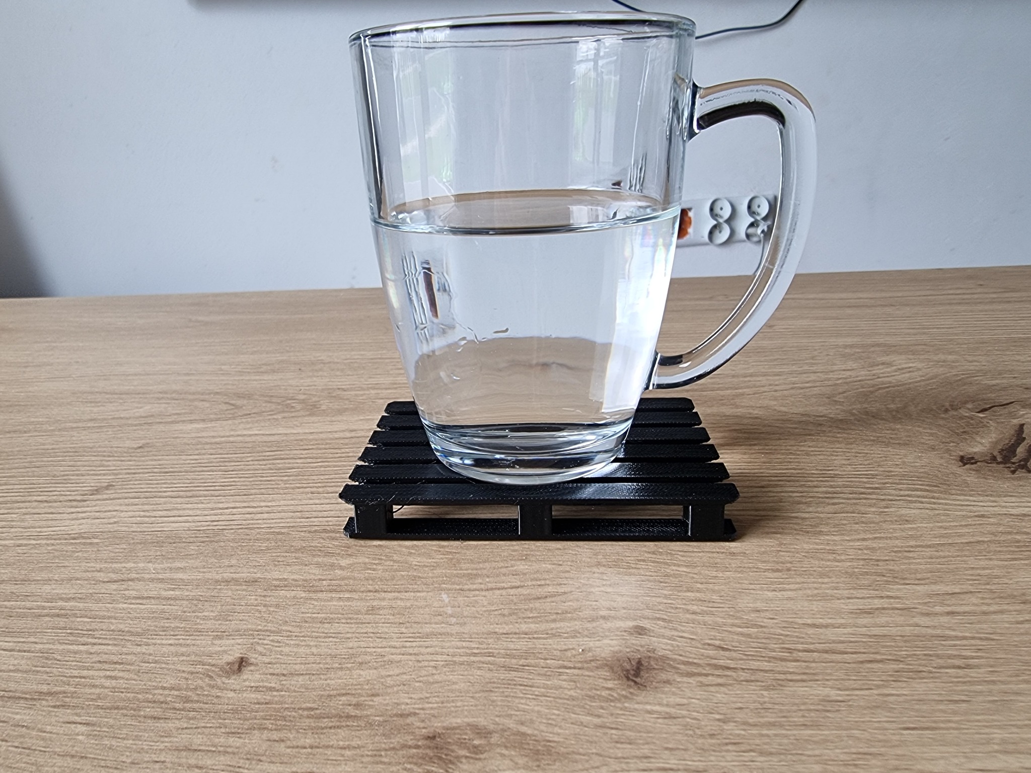 Szklanka z wodą stojąca na czarnej, plastikowej podstawce w kształcie miniaturowej palety na drewnianym blacie biurka. W tle biała ściana i listwa zasilająca.