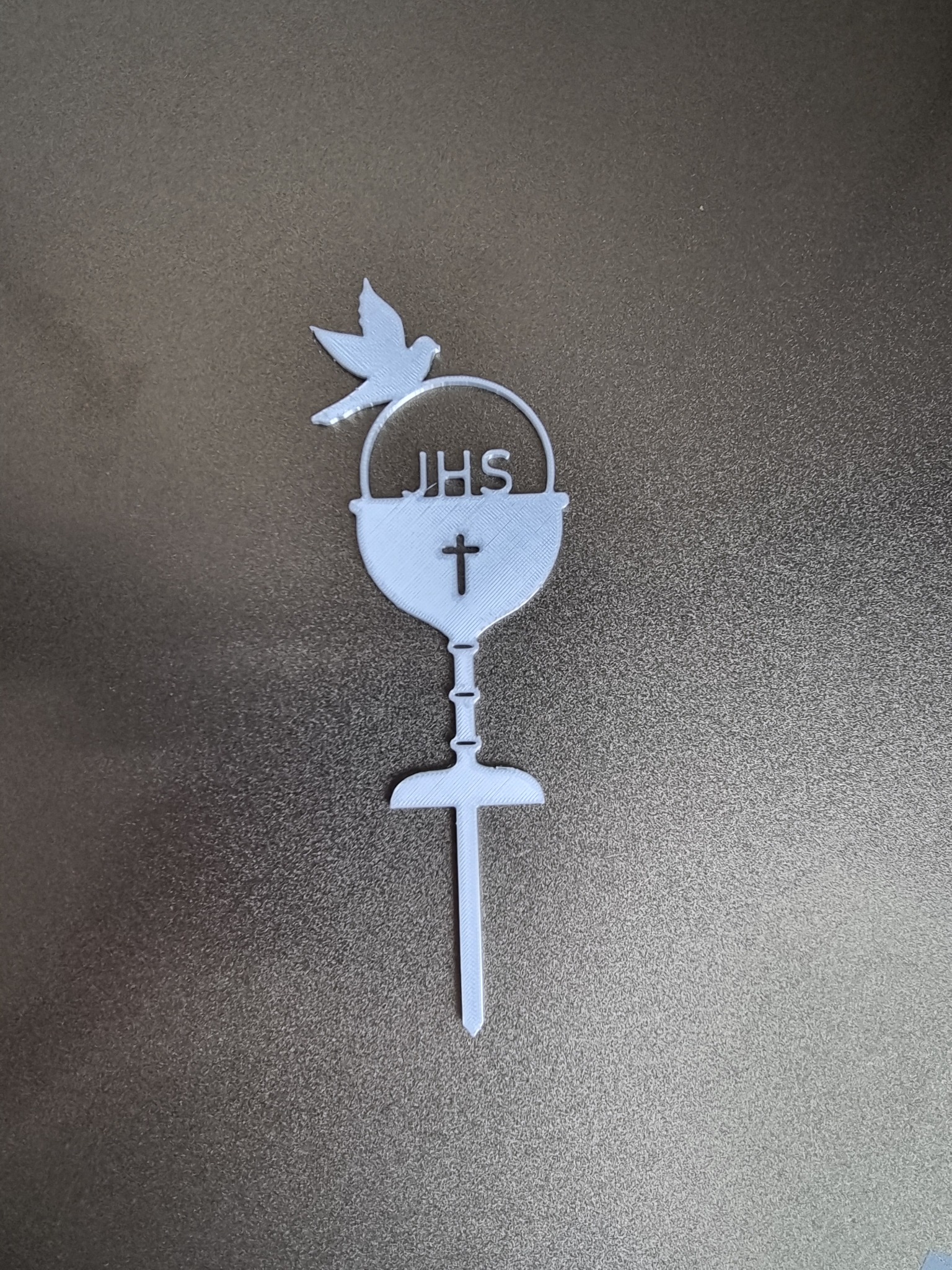 Biała, płaska ozdoba komunijna w kształcie kielicha z hostią, gołębiem i napisem JHS, wykonana prawdopodobnie techniką druku 3D, leżąca na brązowym tle.