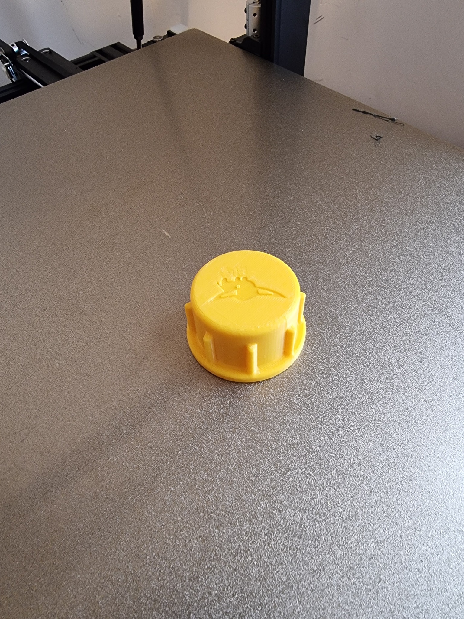 Żółta, okrągła nakładka z wytłoczonym logo na szarej powierzchni drukarki 3D.