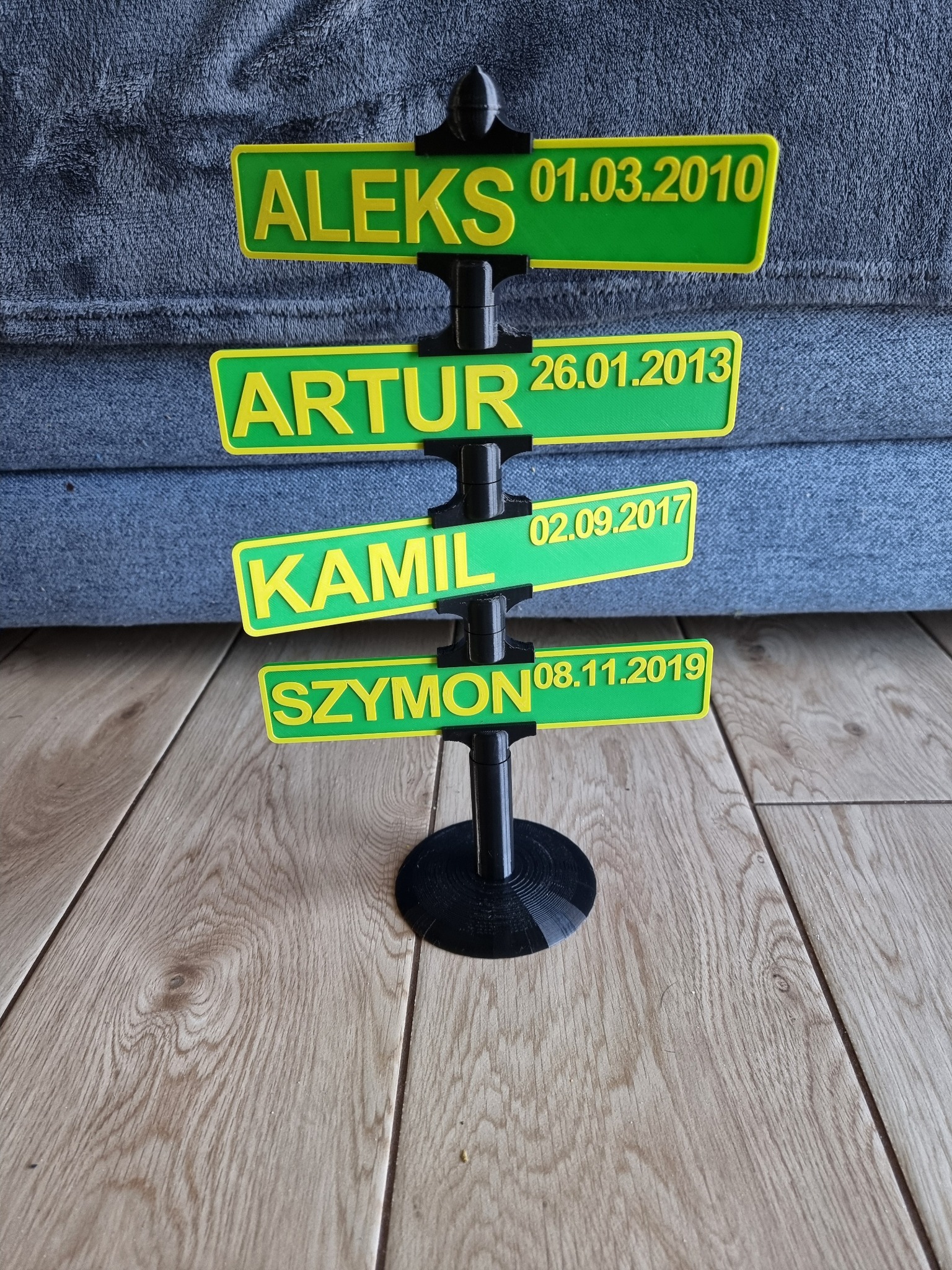Personalizowany drogowskaz z imionami (Aleks, Artur, Kamil, Szymon) i datami urodzenia, wykonany w technologii druku 3D, prezentowany na drewnianej podłodze.