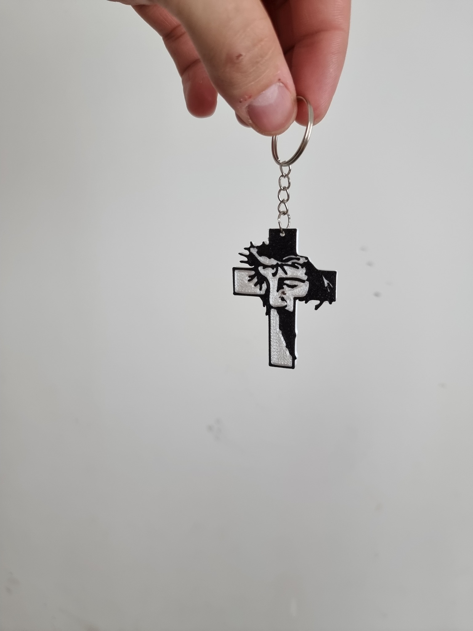 Ręka trzyma brelok z motywem krzyża z wizerunkiem Jezusa, wykonany z czarnego i srebrnego tworzywa, zawieszony na metalowym łańcuszku z kółkiem.