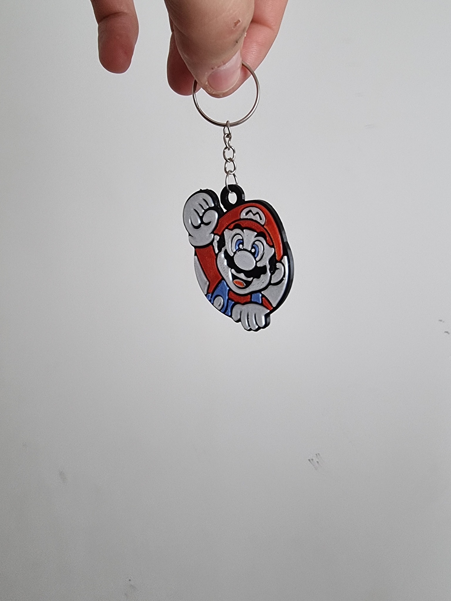 Brelok z postacią Mario w czerwonej czapce, trzymany w palcach na tle białej ściany, wykonany z tworzywa sztucznego z czarnym obramowaniem.