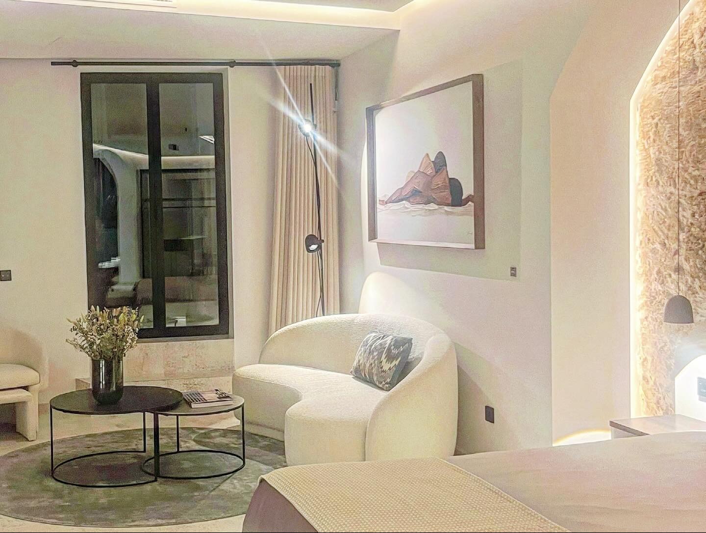 Jasne wnętrze sypialni z okrągłym, białym fotelem, stolikiem kawowym z bukietem suszonych kwiatów i obrazem na ścianie, oświetlone lampą podłogową i lampką nocną.