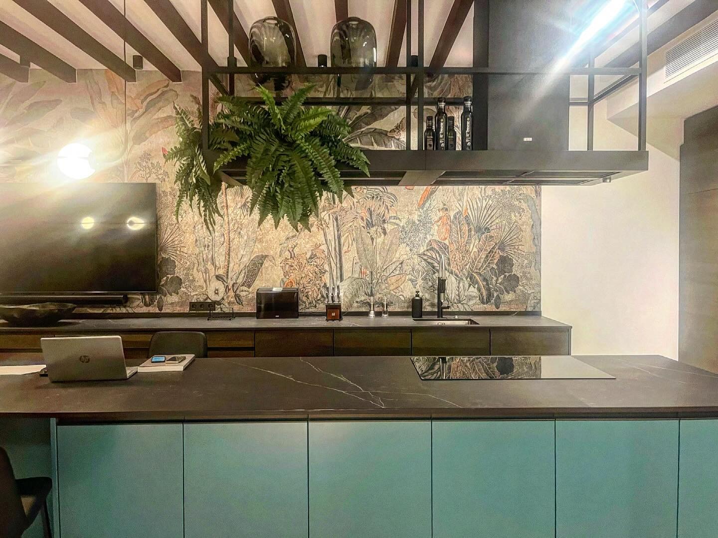 Nowoczesna kuchnia z wyspą, fronty szafek w odcieniu pastelowej zieleni, blat z ciemnego kamienia z wyraźnym użyleniem, ściana wykończona tapetą w motywy roślinne, nad wyspą konstrukcja...