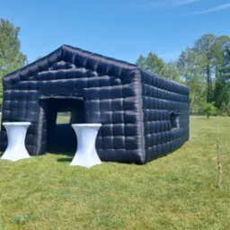 Czarny dmuchany pawilon w kształcie domu z dwoma białymi stolikami koktajlowymi przed wejściem, na trawiastym terenie w słoneczny dzień.