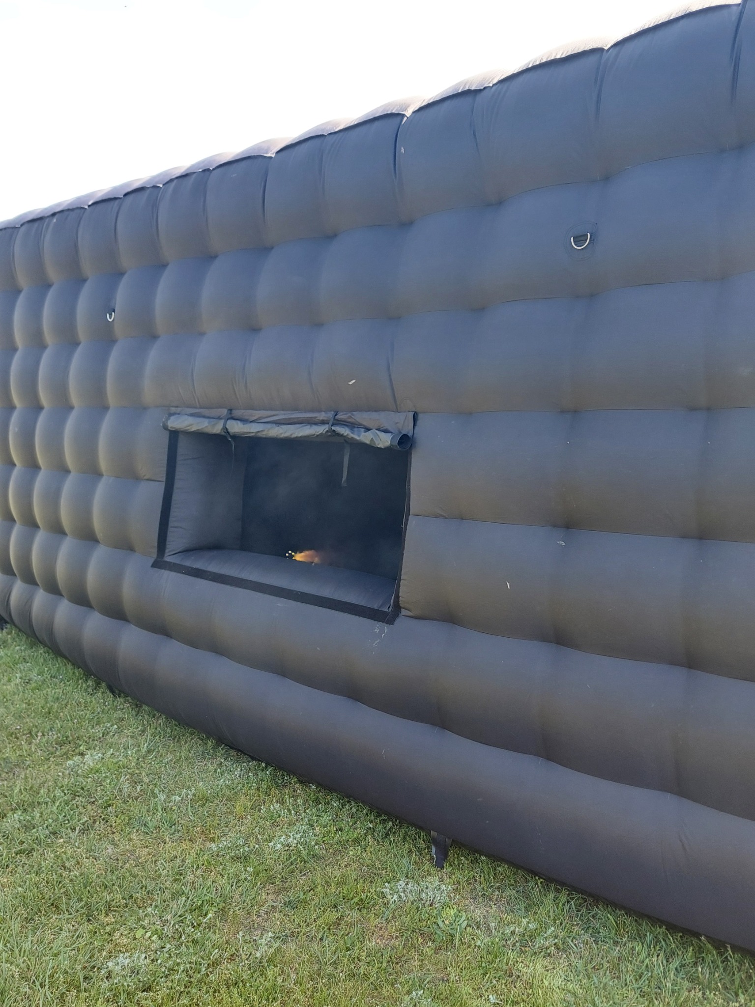 Czarna, dmuchana konstrukcja z oknem, ustawiona na trawie. Wewnątrz okna widoczne światła.