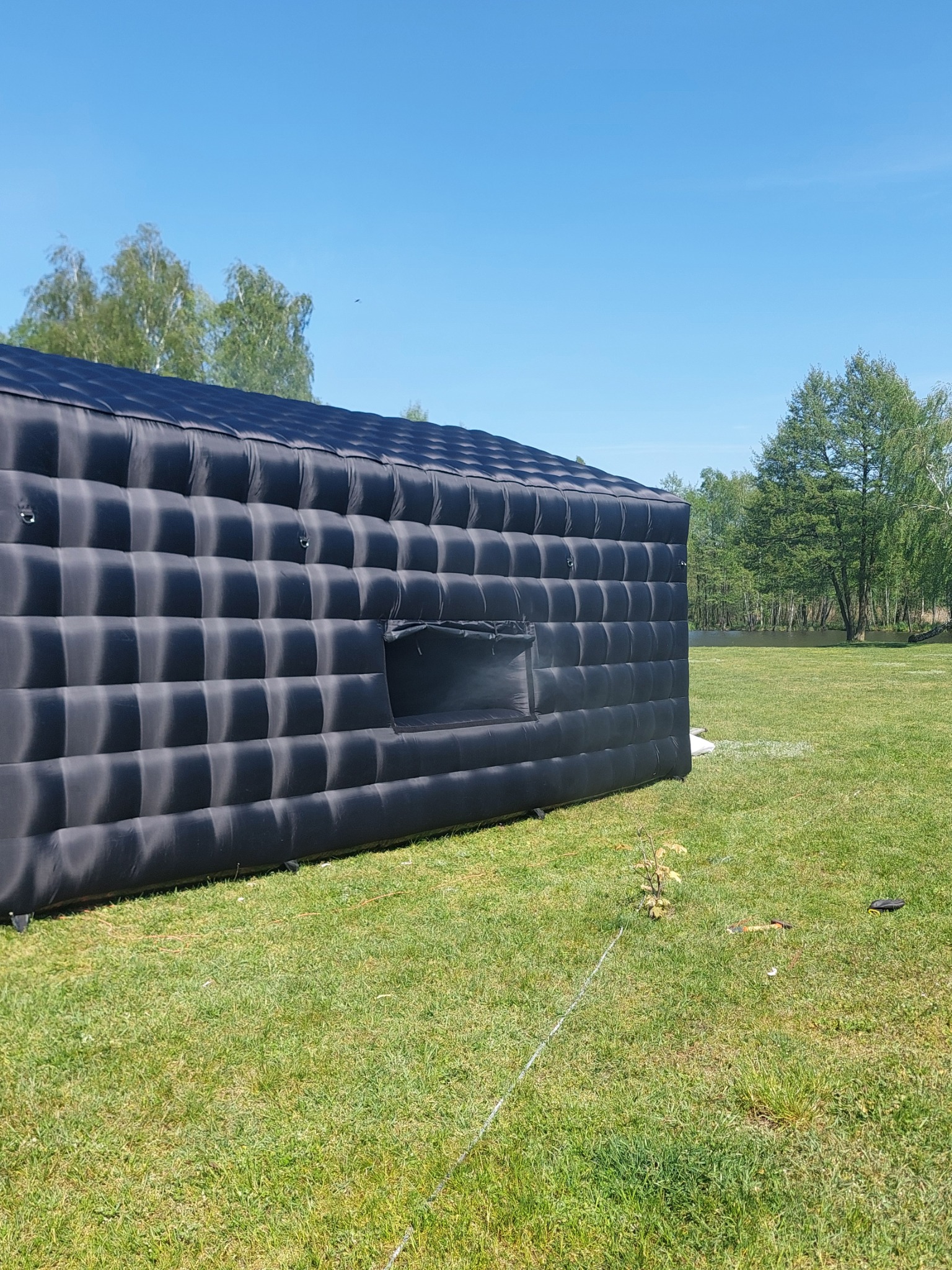 Czarna, dmuchana konstrukcja w kształcie budynku z oknem, ustawiona na trawniku w słoneczny dzień. Widoczne drzewa w tle.