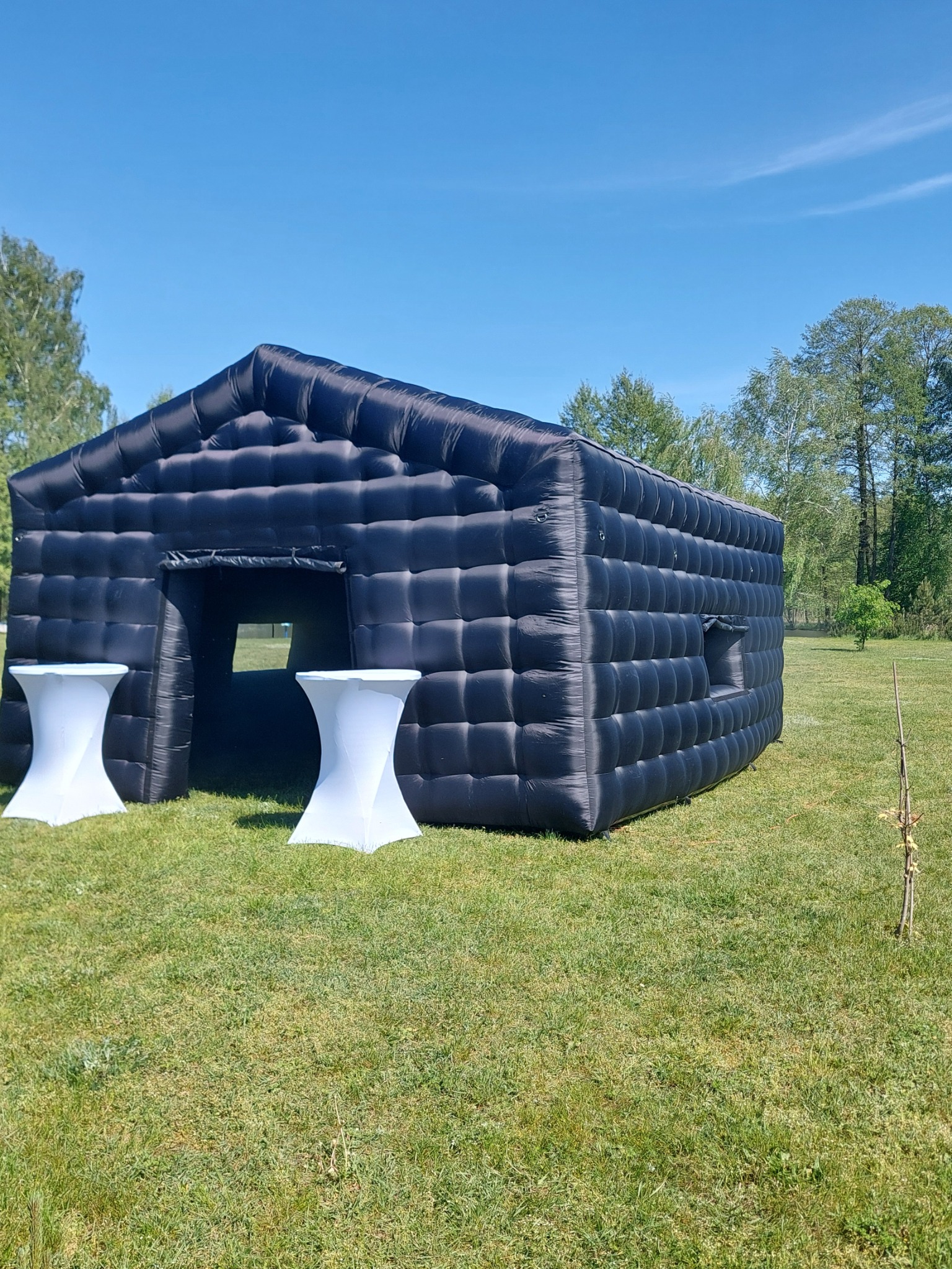 Czarny dmuchany pawilon w kształcie domu z dwoma białymi stolikami koktajlowymi przed wejściem, na trawiastym terenie w słoneczny dzień.