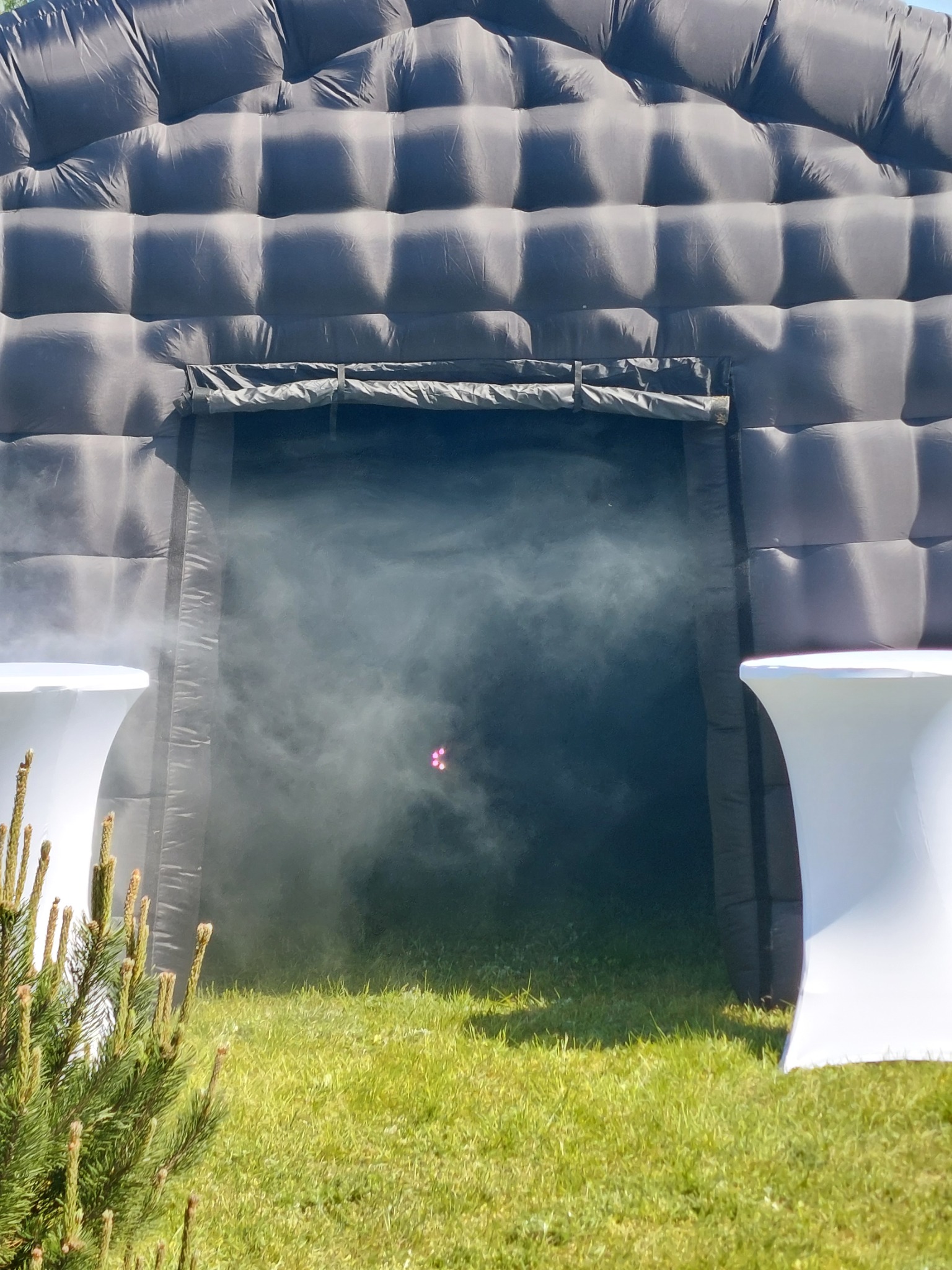 Wejście do ciemnego, dmuchanego namiotu z efektem mgły, widoczne dwa białe stoliki po bokach i zielona trawa.