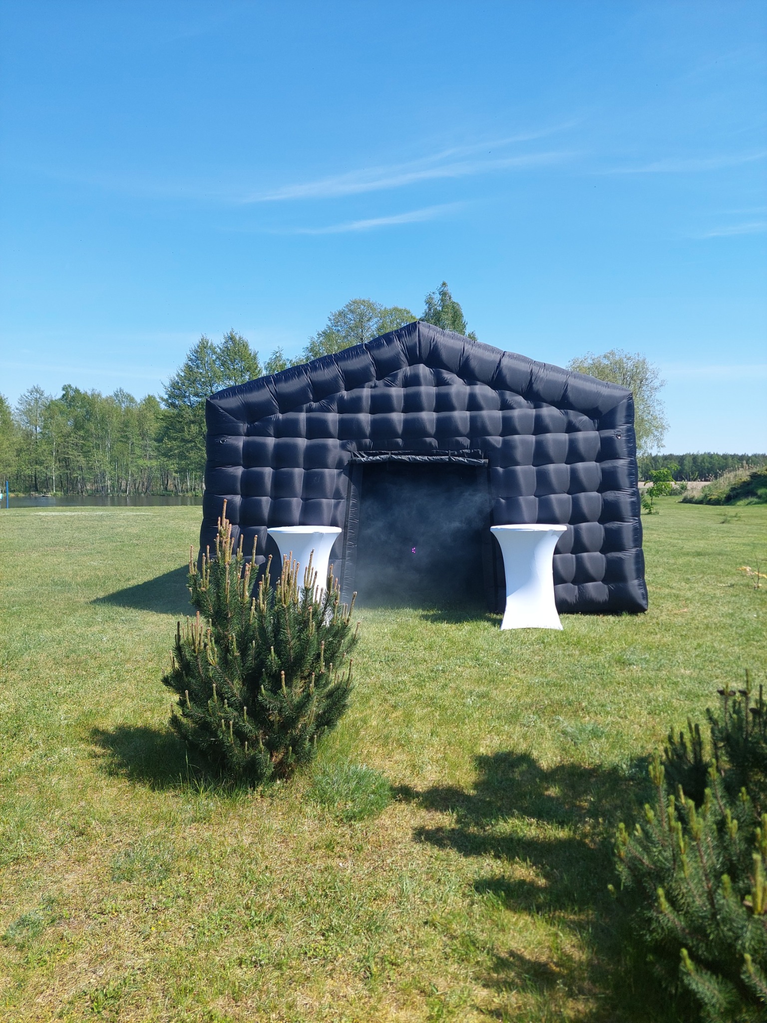 Czarna, dmuchana konstrukcja w kształcie domu na zielonym trawniku, z dwoma białymi stolikami koktajlowymi po bokach wejścia, otoczona mgłą, na tle drzew i błękitnego nieba.