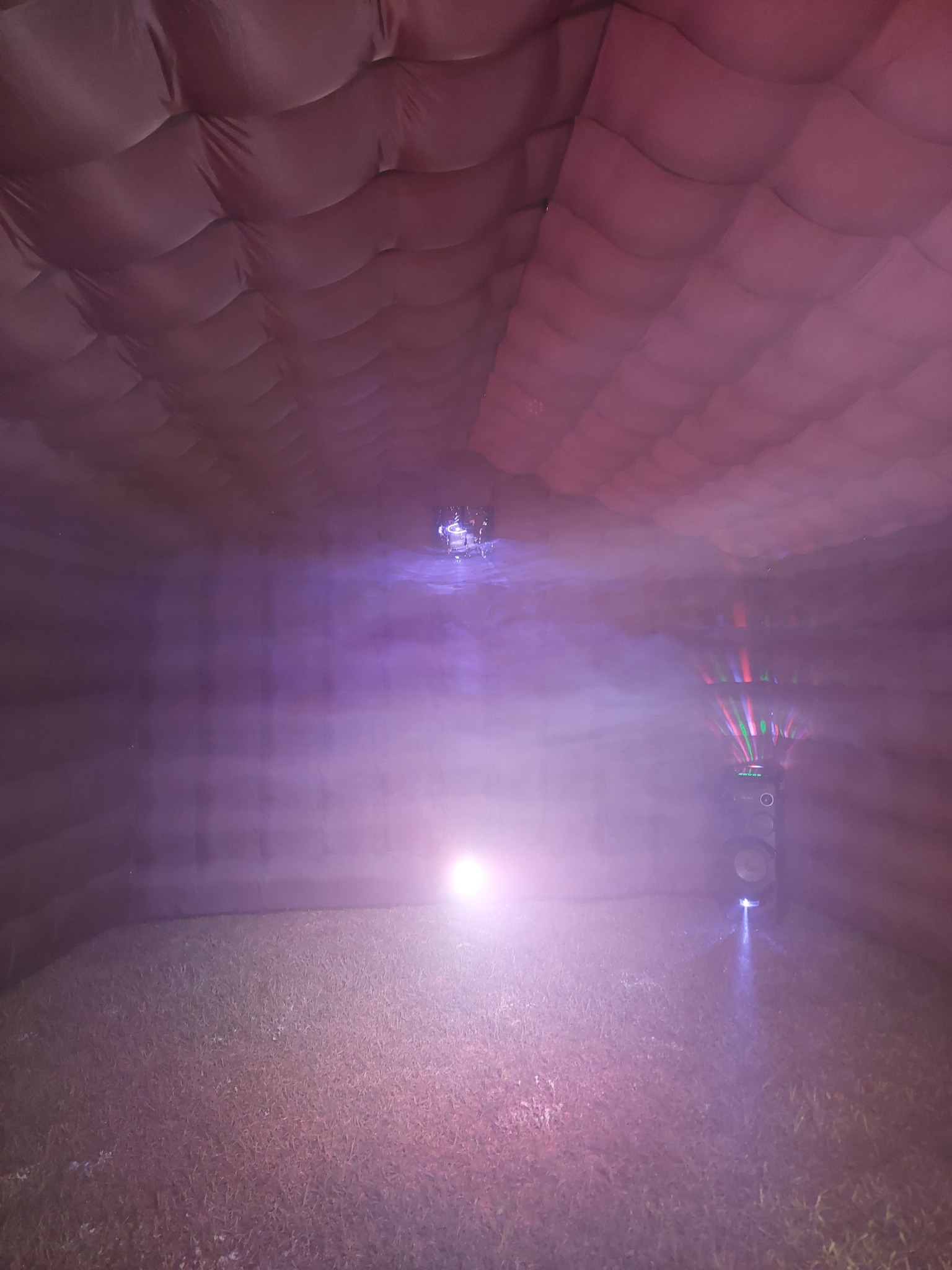 Wnętrze ciemnego dmuchanego namiotu z podłogą pokrytą sztuczną trawą, oświetlone przez reflektor i kolumnę głośnikową z efektami świetlnymi.