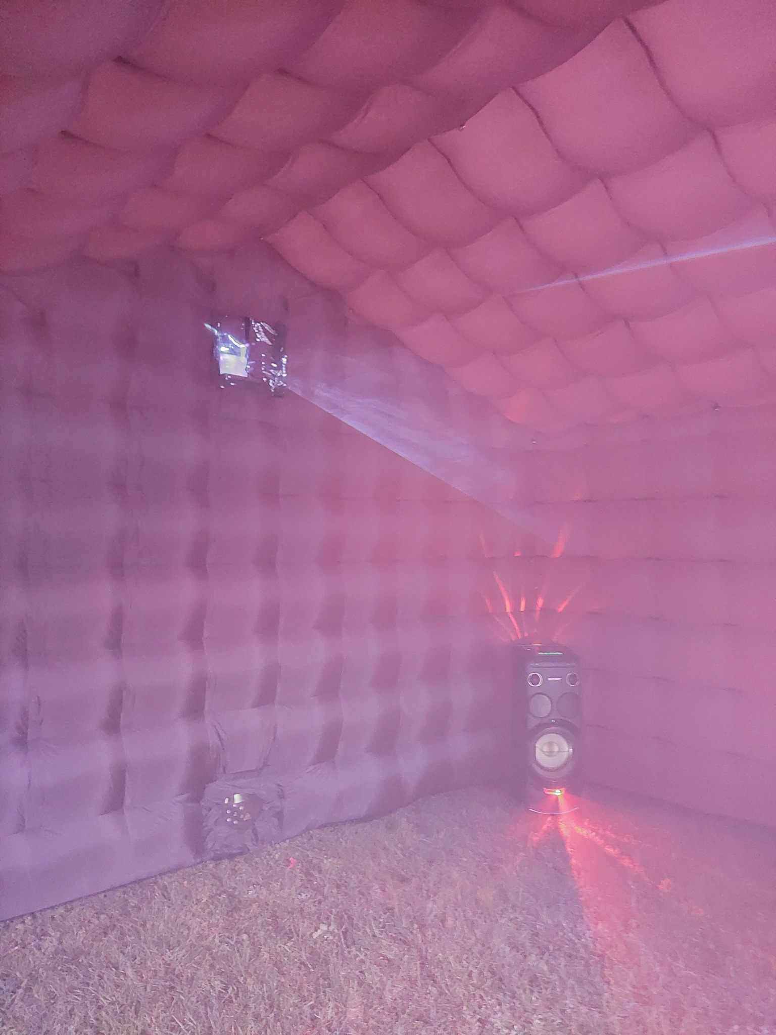 Wnętrze dmuchanej konstrukcji oświetlone różowym światłem, z widocznym projektorem i głośnikiem na tle trawy.