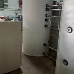 Dwa białe, pionowe zbiorniki buforowe ciepła w kotłowni, z podłączonymi rurami instalacji hydraulicznej, widoczne elementy armatury i osprzętu, w tle ściana z instalacjami.