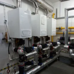Trzy identyczne, białe piece gazowe zamontowane na ścianie z szarą glazurą, z widocznymi rurami i zaworami, izolowanymi folią aluminiową, w pomieszczeniu technicznym.
