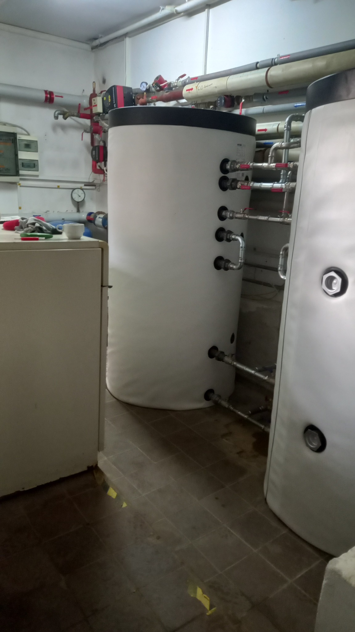 Dwa białe, pionowe zbiorniki buforowe ciepła w kotłowni, z podłączonymi rurami instalacji hydraulicznej, widoczne elementy armatury i osprzętu, w tle ściana z instalacjami.