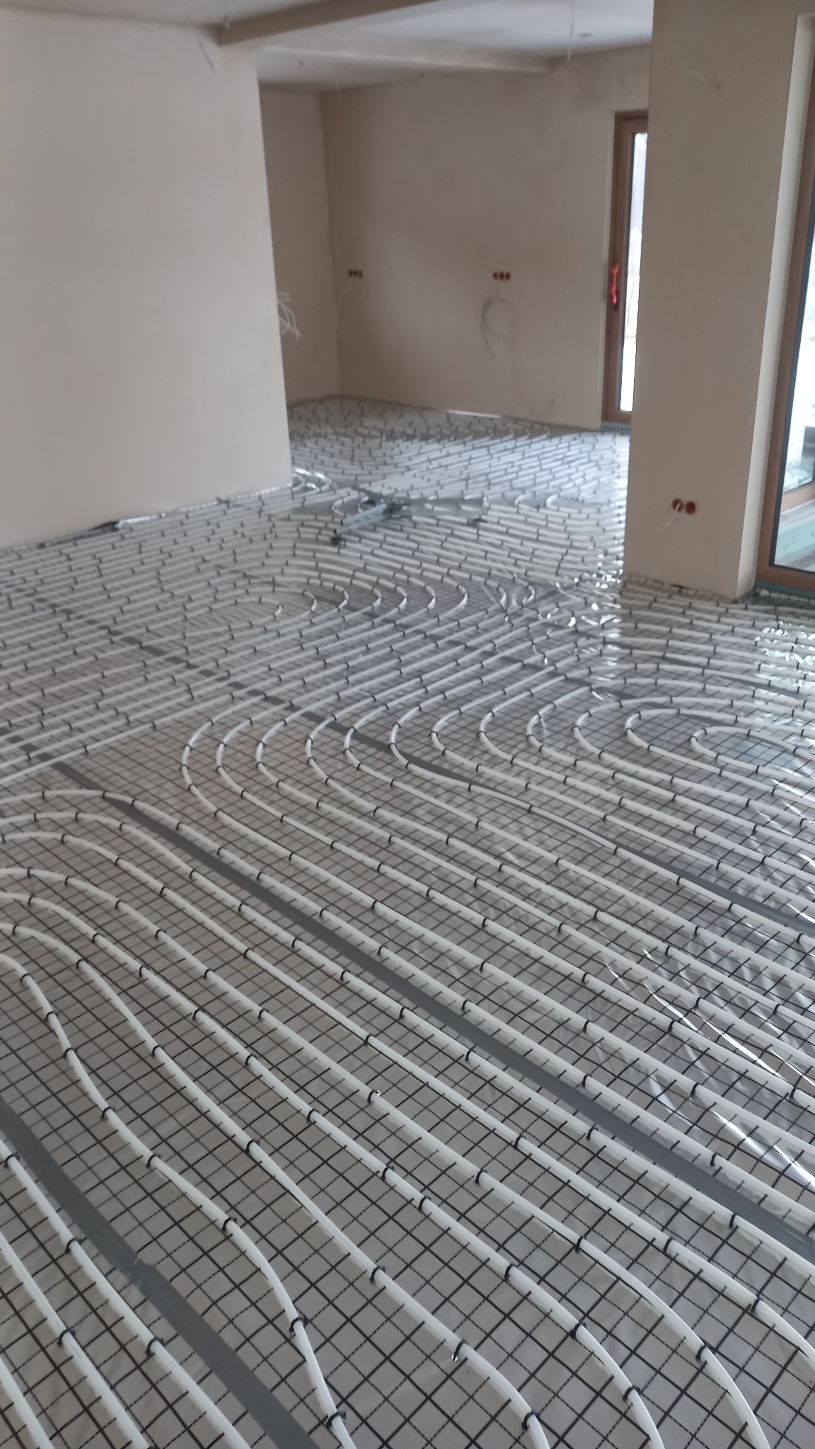 Rozłożona instalacja ogrzewania podłogowego z białych rurek na siatce, widok z góry w surowym wnętrzu.