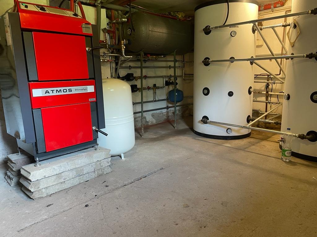 Czerwono-czarny piec marki Atmos na betonowych blokach, obok biały zbiornik wyrównawczy i dwa białe akumulatory ciepła z rurami, w piwnicy.
