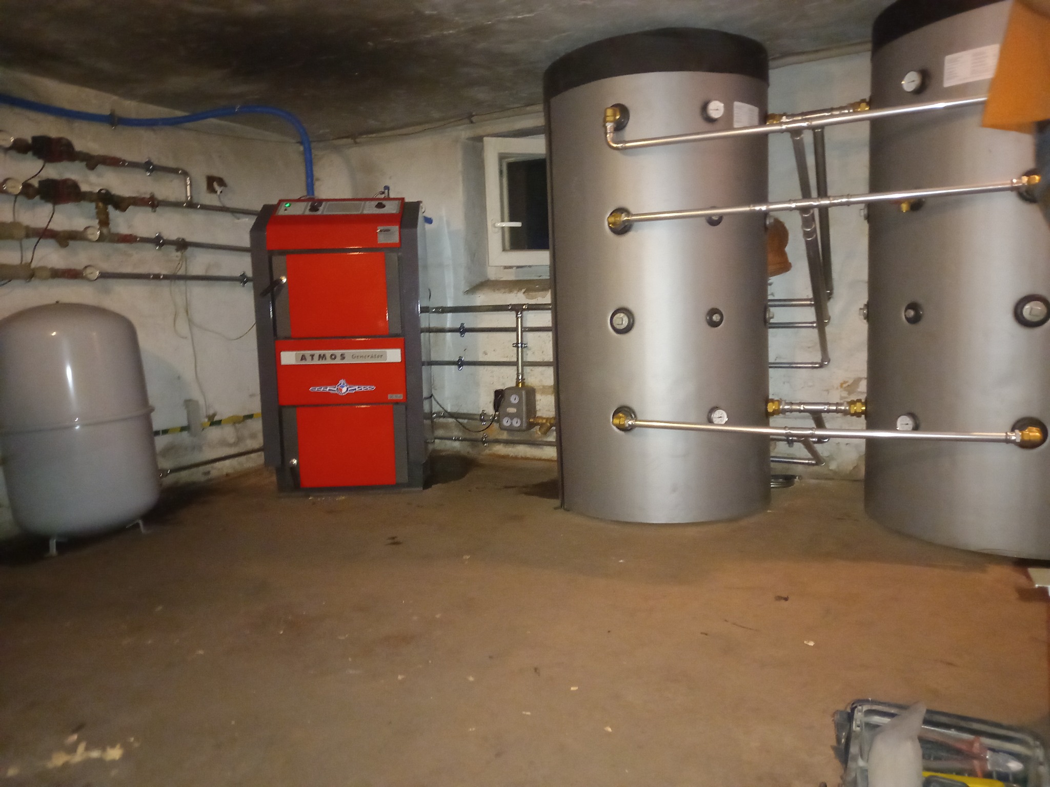 Czerwony piec grzewczy Atmos Generator, dwa srebrne zbiorniki akumulacyjne z rurami, szara instalacja hydrauliczna na ścianie w piwnicy z małym oknem.