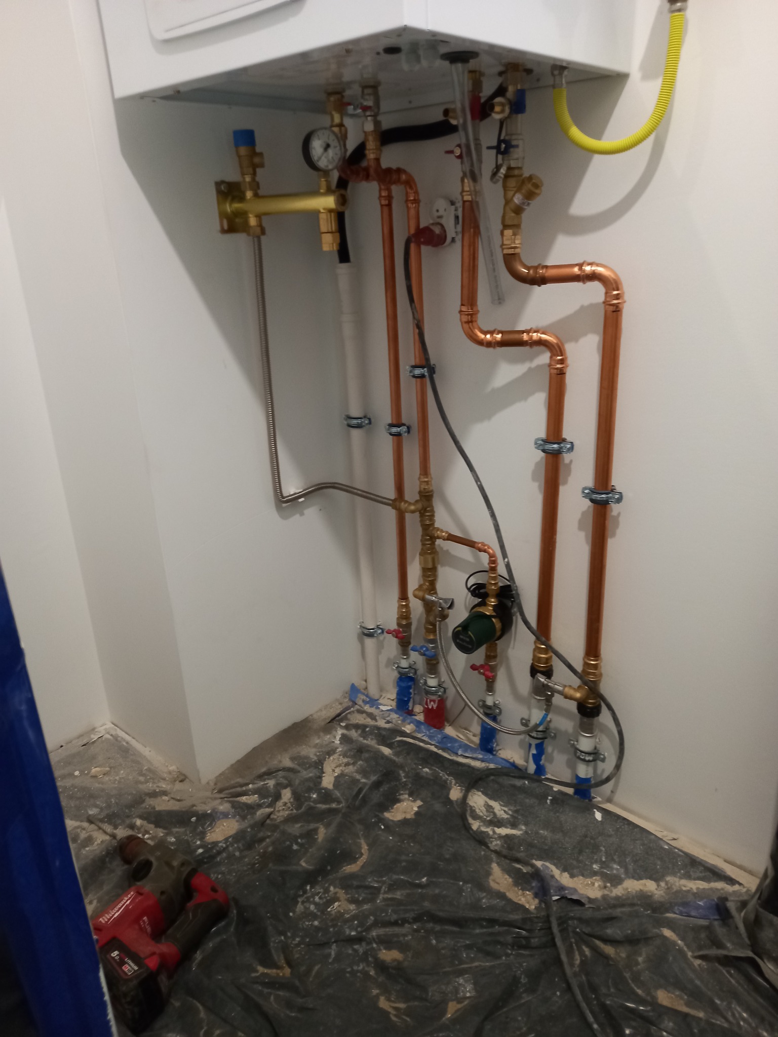 Świeżo zamontowany piec gazowy z miedzianą instalacją, widoczne rury, zawory i manometr, na podłodze folia ochronna i wiertarka.